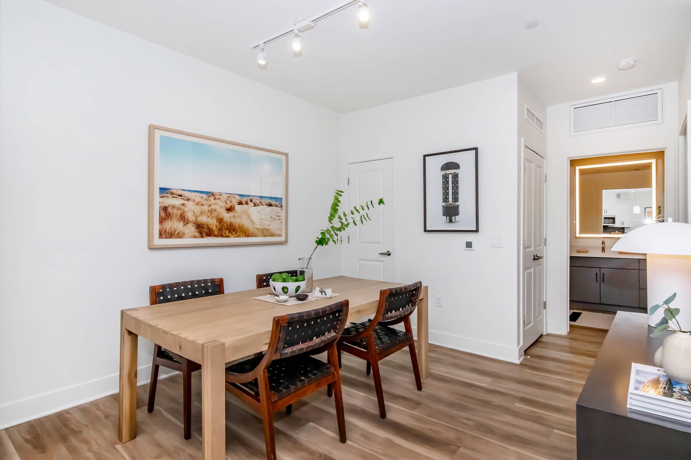 A modern dining area featuring a wooden table with four black woven chairs. A large framed landscape photograph of a beach scene hangs on the wall. A green plant centerpiece is placed on the table, and there is a contemporary lamp and framed artwork on a nearby console. Soft lighting enhances the bright, minimalist decor.