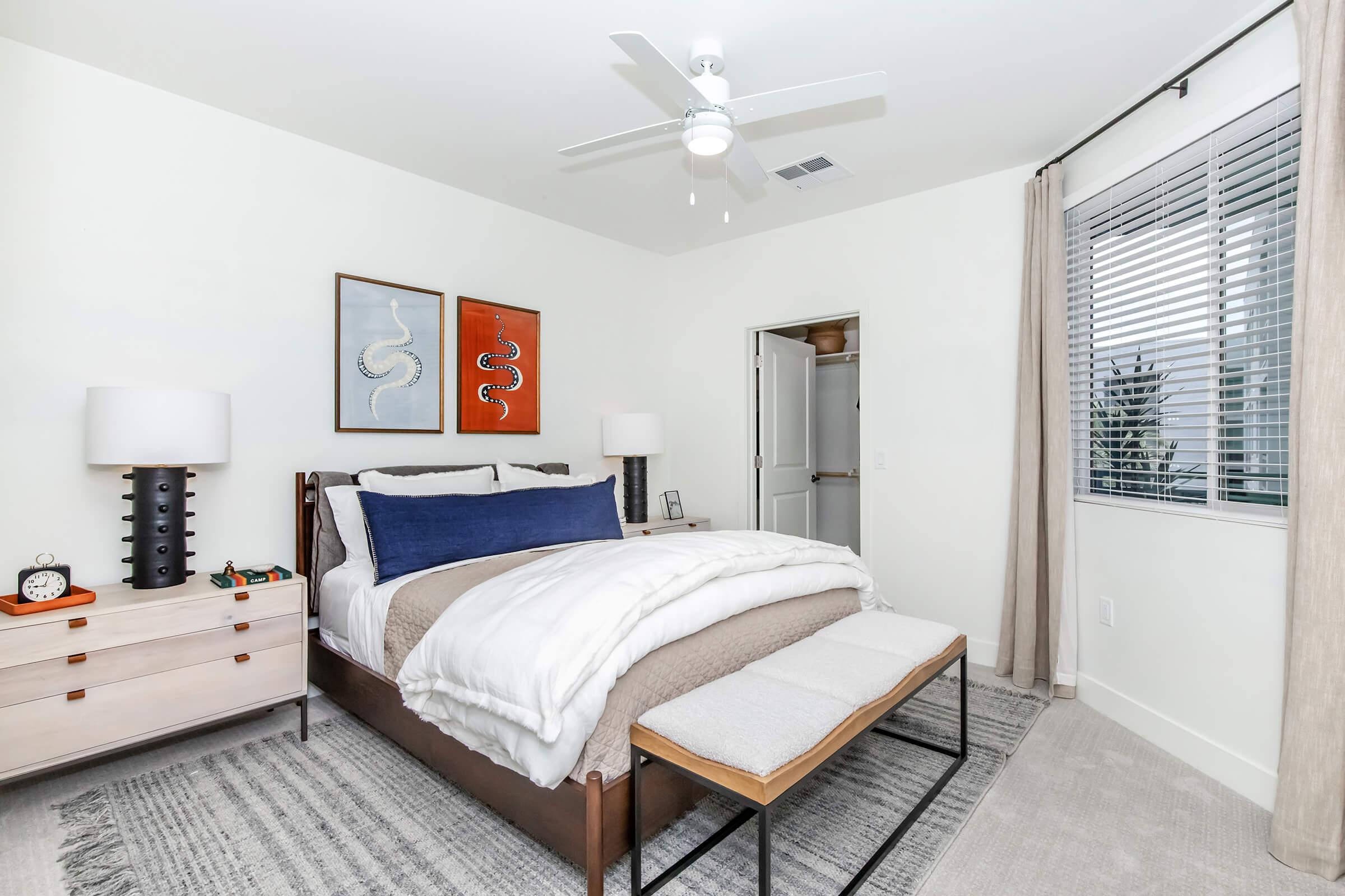 A modern bedroom featuring a wooden bed with a white comforter and blue accent pillows. There are two bedside lamps, abstract artwork on the walls, and a light-colored rug. A ceiling fan is visible, and a window with blinds allows natural light into the space.