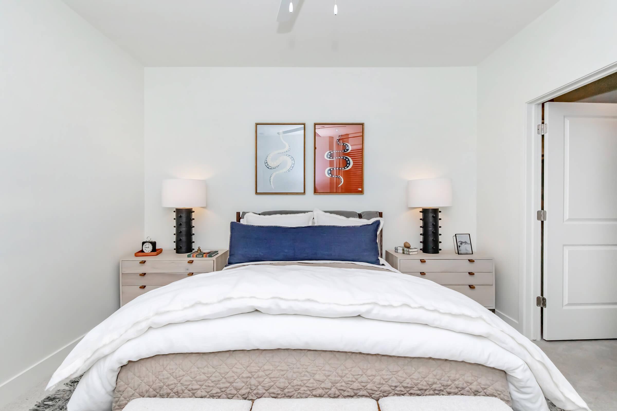 A modern bedroom featuring a neatly made bed with a white comforter and a navy blue accent pillow. On either side of the bed, there are stylish nightstands with lamps. Above the bed, two abstract framed prints hang on the wall. The room has light-colored walls and a door partially open, revealing a glimpse of the hallway.