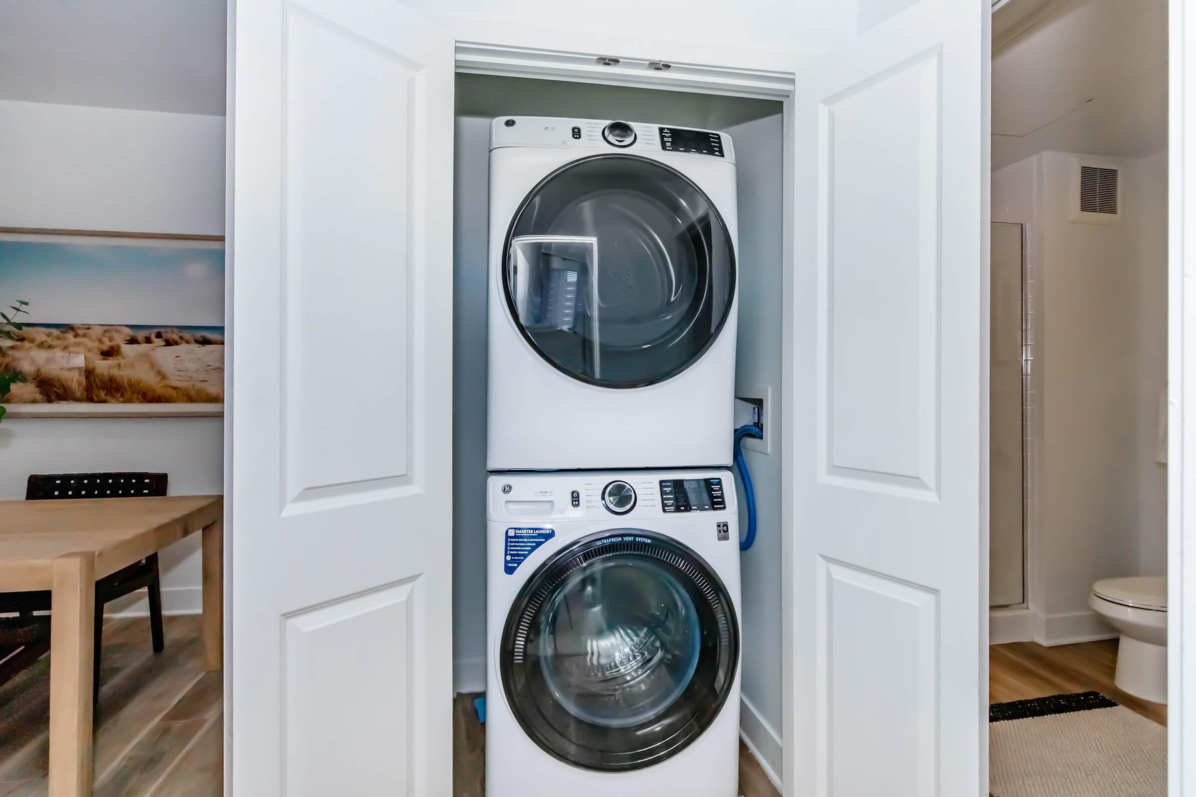 A stackable washer and dryer unit installed in a laundry closet, with white cabinetry and a framed beach scene visible in the background. A dining table and chairs are positioned nearby, and a bathroom is partially seen to the right.