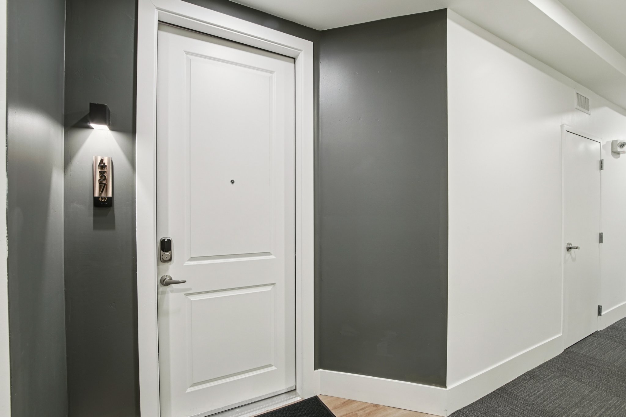 A closed white door with a small peephole, framed by a dark gray wall. A modern light fixture is mounted next to the door, providing illumination. The area features a light-colored floor and a minimalistic design, with an adjacent white door partly visible.