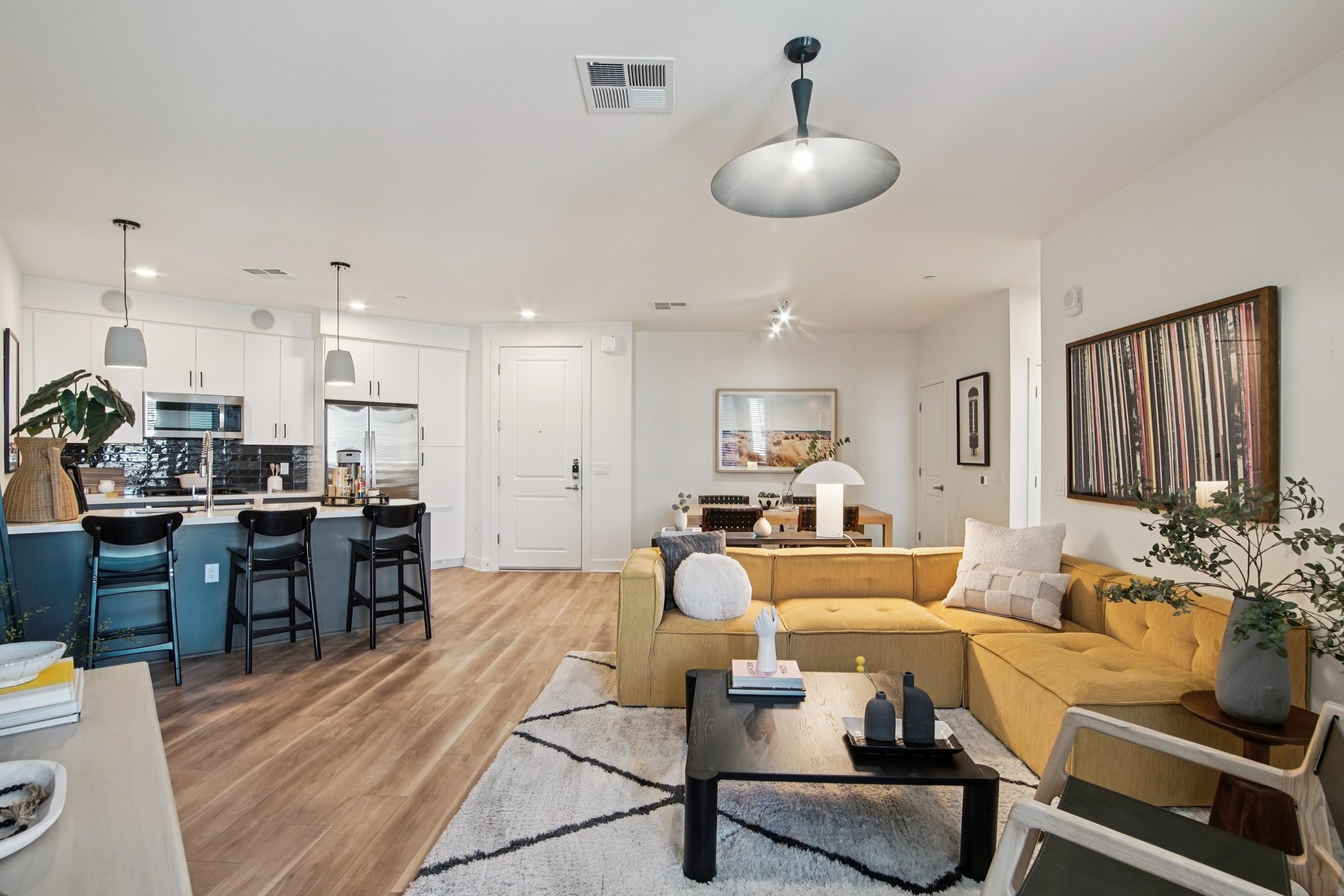 A modern living room with a yellow sectional sofa, a black coffee table, and decorative items. In the background, a kitchen with white cabinets and stainless steel appliances, bar stools at the counter, and a dining area. Natural light fills the space, accented by artwork on the walls.