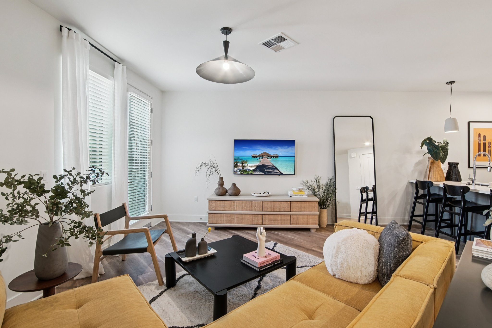 A modern living room with a yellow sofa and a black coffee table. There’s a flat-screen TV on a wooden console showcasing a beach scene. Large windows with sheer curtains allow natural light. A mirror and stylish décor, including plants and artworks, enhance the inviting atmosphere.