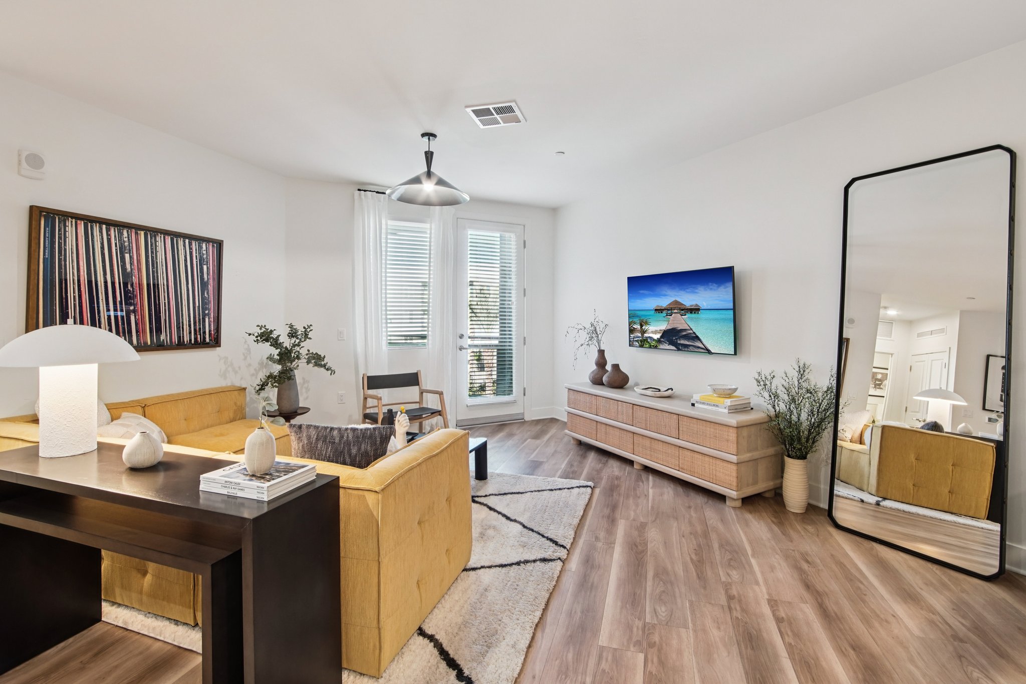 Modern living room featuring a yellow sofa, stylish wooden furniture, a large mirror, and a flat-screen TV displaying a beach scene. Natural light streams in through large windows with sheer curtains, and decorative plants add a fresh touch to the interior. A contemporary artwork hangs on the wall.