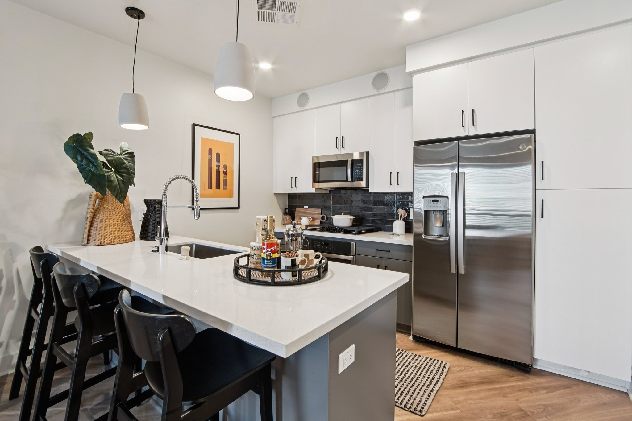 Modern kitchen featuring stainless steel appliances, a sleek island with bar stools, and a stylish decor. The space includes white cabinets, dark accents, and complementary artwork on the walls, creating a bright and inviting atmosphere ideal for cooking and entertaining.