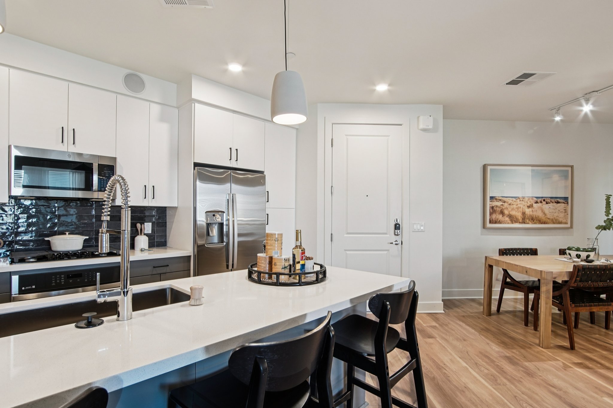 Modern kitchen with white cabinetry, stainless steel appliances, and a sleek countertop. A circular tray with drinks is on the island, featuring barstools. A dining area with a wooden table and black chairs is visible, along with a framed landscape picture on the wall. Natural light fills the space.