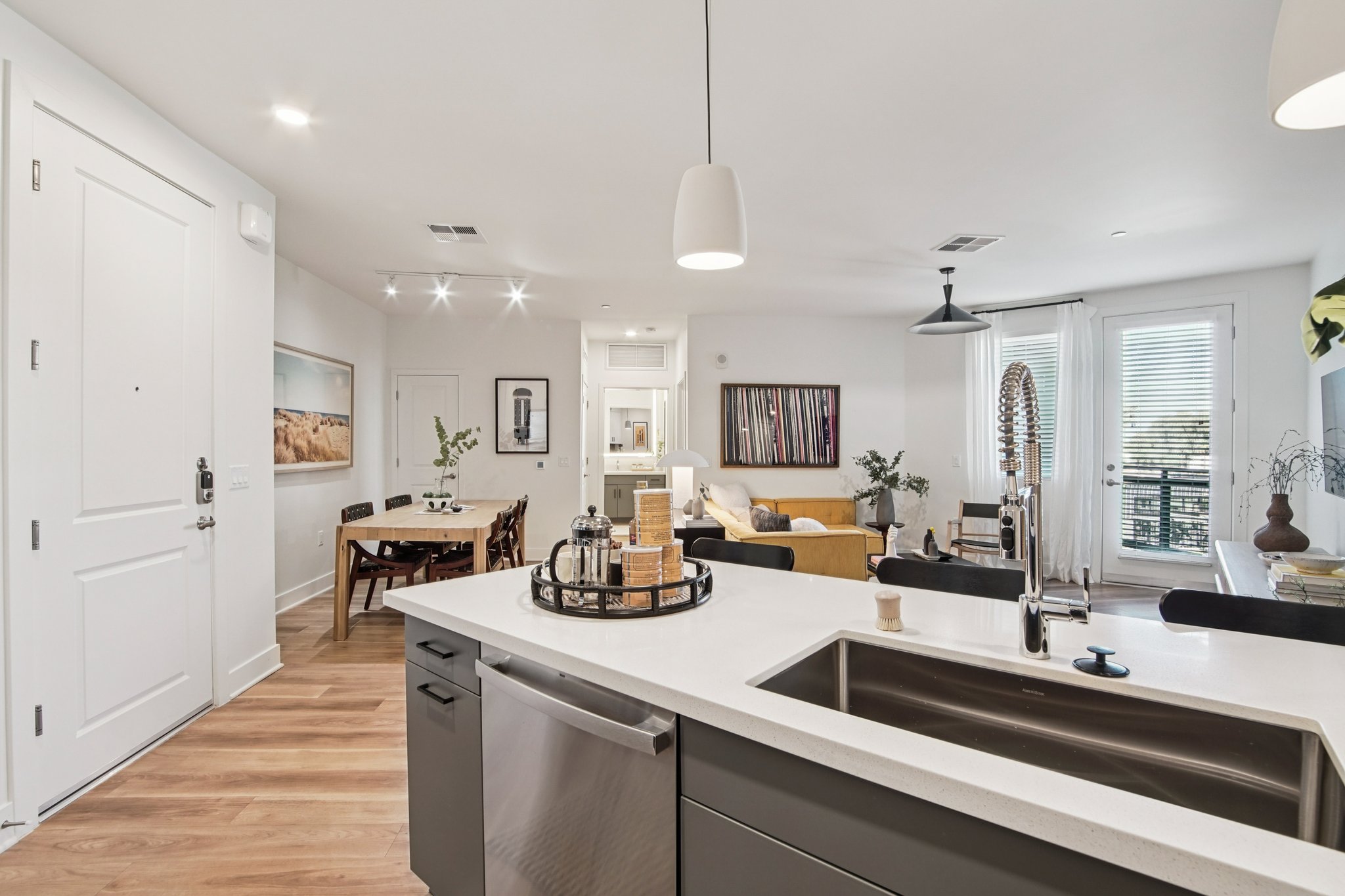 Modern open-concept living space featuring a stylish kitchen with a central island, stainless steel appliances, and a dining area with a round table. The living room has a comfortable sofa, decorative plants, and large windows that provide natural light, creating a warm and inviting atmosphere.