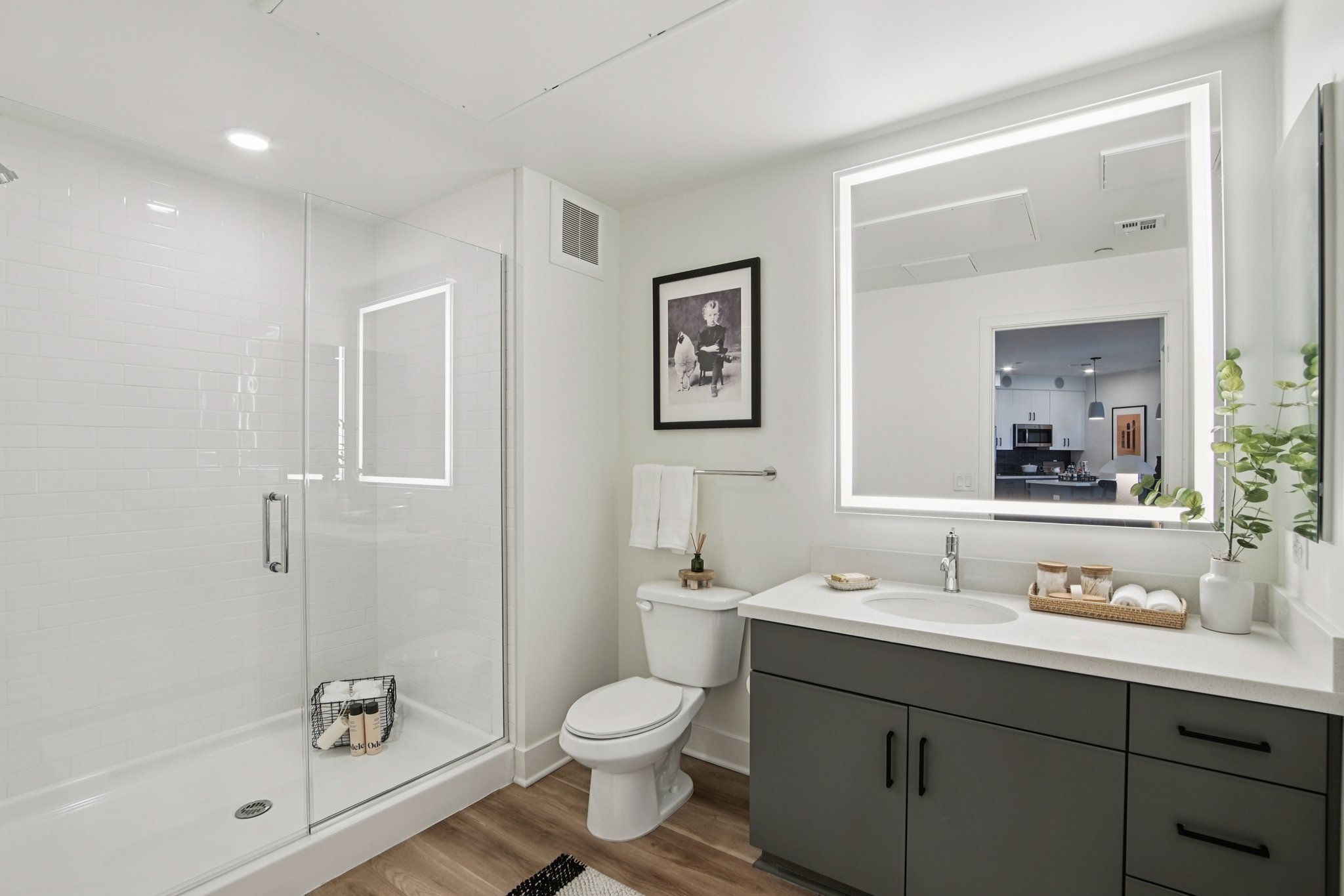 Modern bathroom featuring a walk-in shower with glass doors, a white toilet, and a double-sink vanity with dark gray cabinetry. Soft lighting is provided by a mirror with built-in illumination. Clean design elements include a framed photo on the wall, decorative towels, and a plant on the countertop.