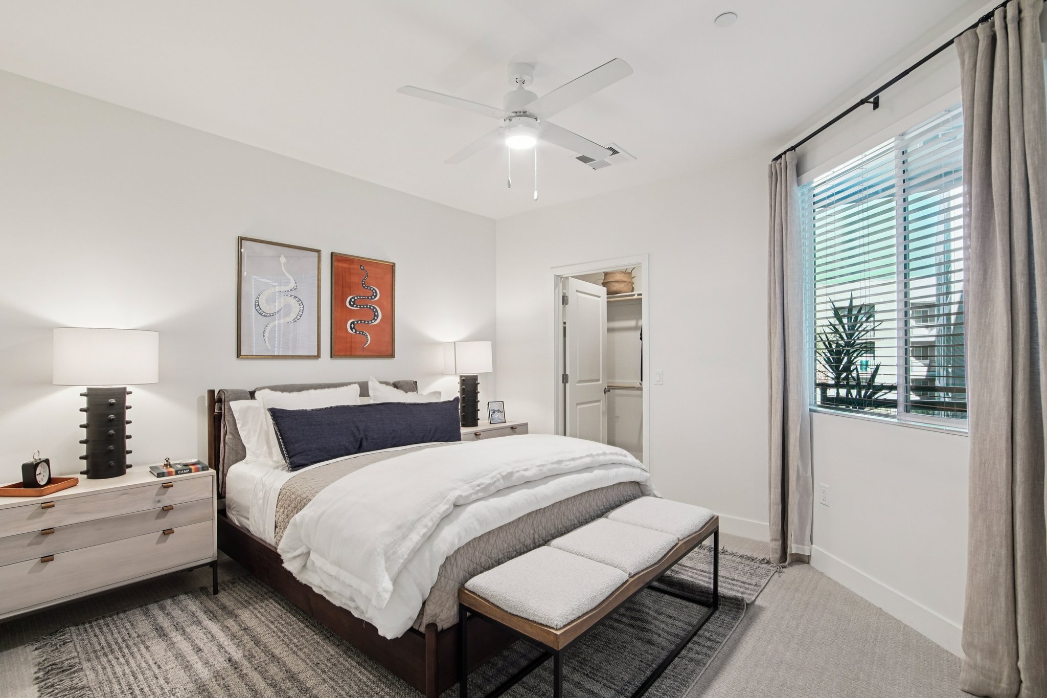 A modern bedroom featuring a king-sized bed with white and gray bedding, two bedside lamps, and a decorative bench at the foot of the bed. The room has light-colored walls, a ceiling fan, and a large window with blinds, allowing natural light to enter. Artwork adorns the walls, enhancing the contemporary design.
