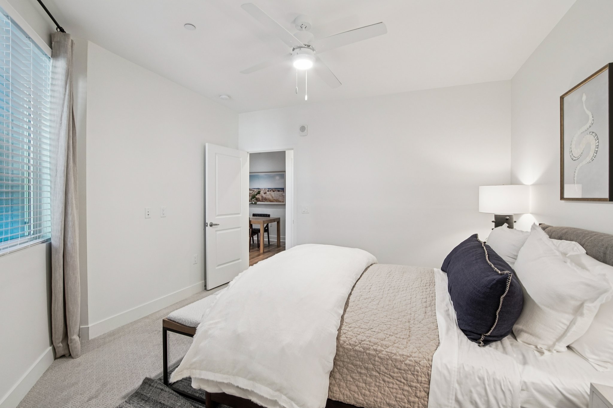 A modern bedroom featuring a neatly made bed with white and navy bedding, a bedside table with a lamp, and a decorative wall art piece. Natural light streams in through a window, and a door leads to another room in the background. The walls are painted white, and the flooring is carpeted.