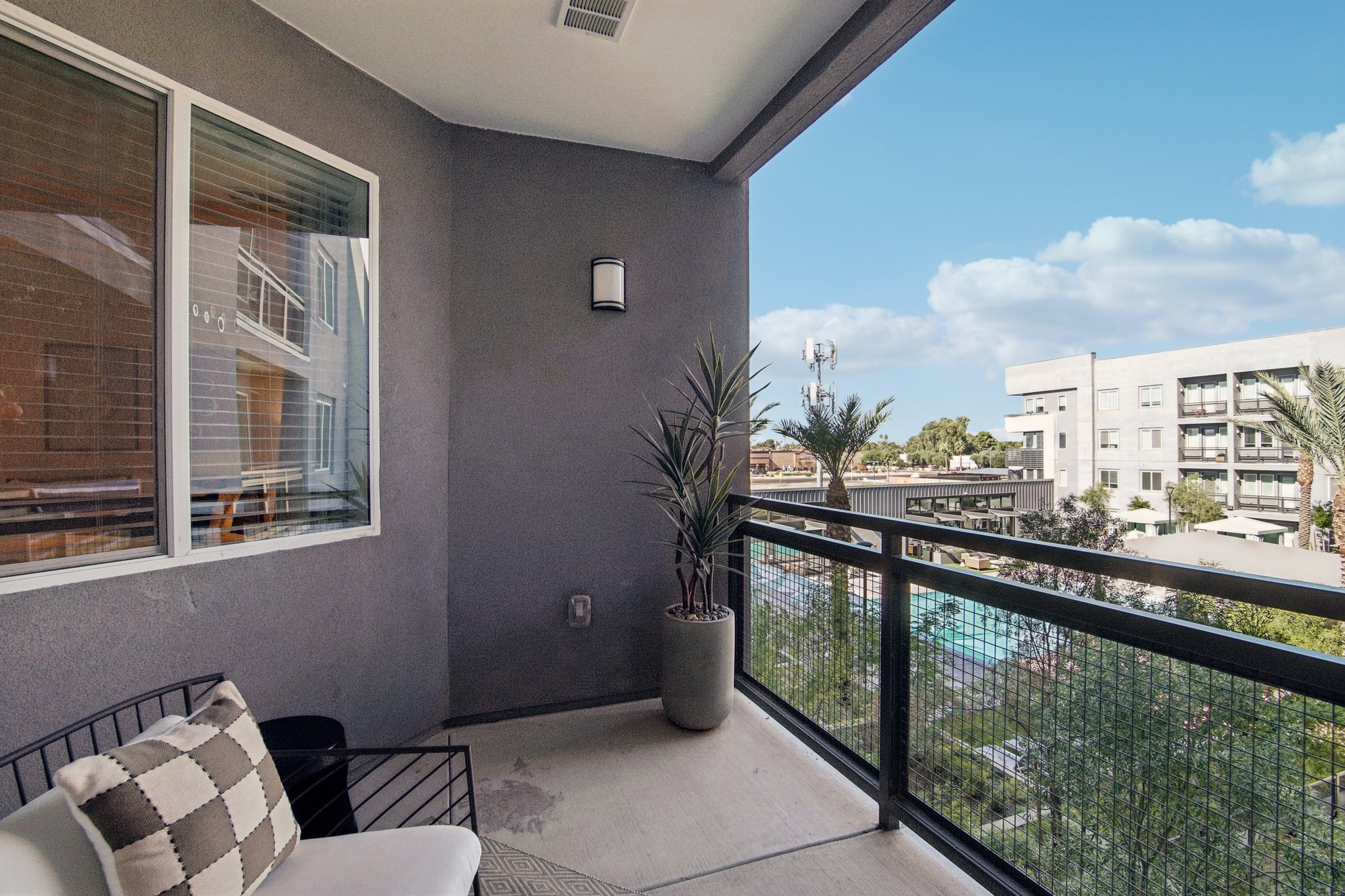 Balcony view featuring a comfortable chair and potted plant, overlooking a landscaped area with a swimming pool and nearby buildings under a blue sky with scattered clouds.