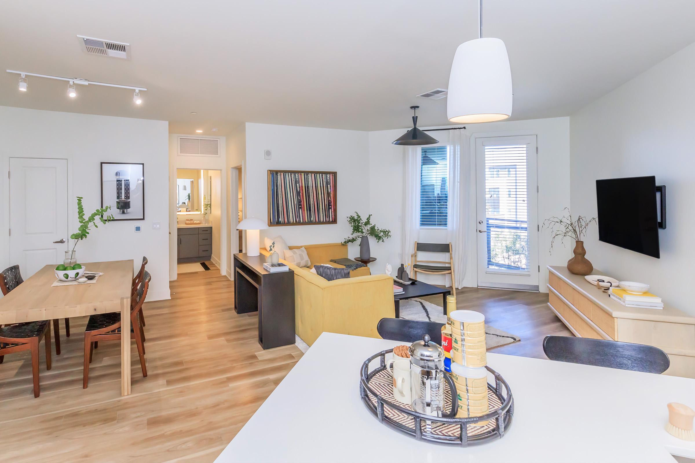 A modern living space featuring a dining area with a wooden table and chairs, a cozy sitting area with a yellow couch, and a sleek entertainment center. Natural light flows through large windows, highlighting minimalist decor and plants. The open layout connects the kitchen and living areas seamlessly.