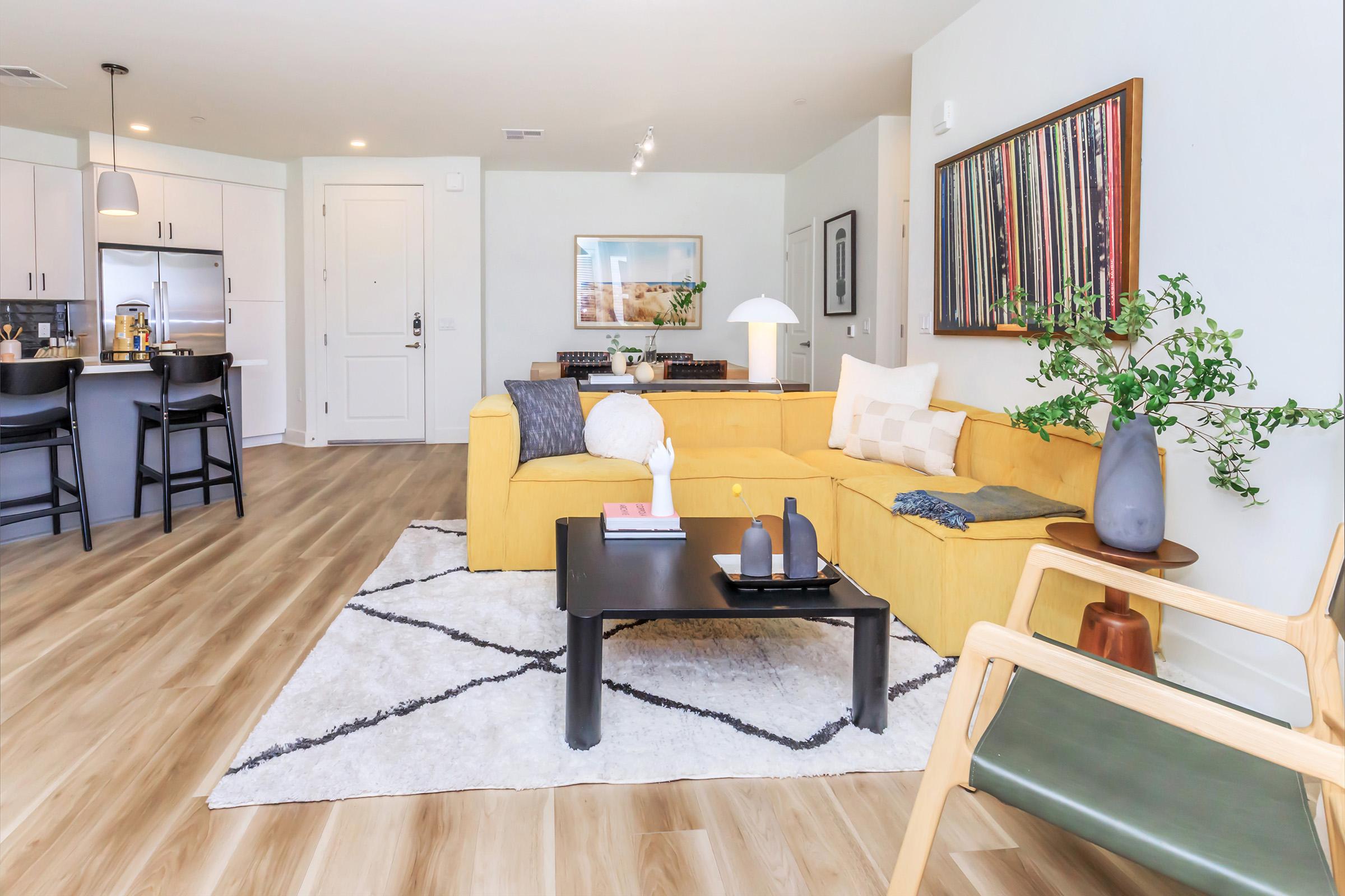 Modern living room featuring a yellow sectional sofa, a black coffee table with decorative items, and a green chair. Natural light fills the space, which includes a rug with a geometric pattern. In the background, there's a kitchen with bar stools and a framed artwork on the wall.