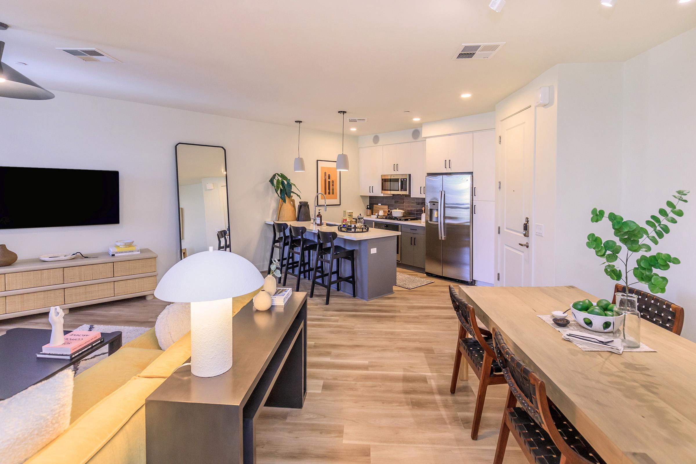Bright and modern living space featuring a kitchen with stainless steel appliances, a dining area with a wooden table and chairs, and a cozy lounge with a yellow sofa and black accents. Large windows allow natural light, and decorative plants add a touch of greenery. A wall-mounted TV enhances the entertainment area.
