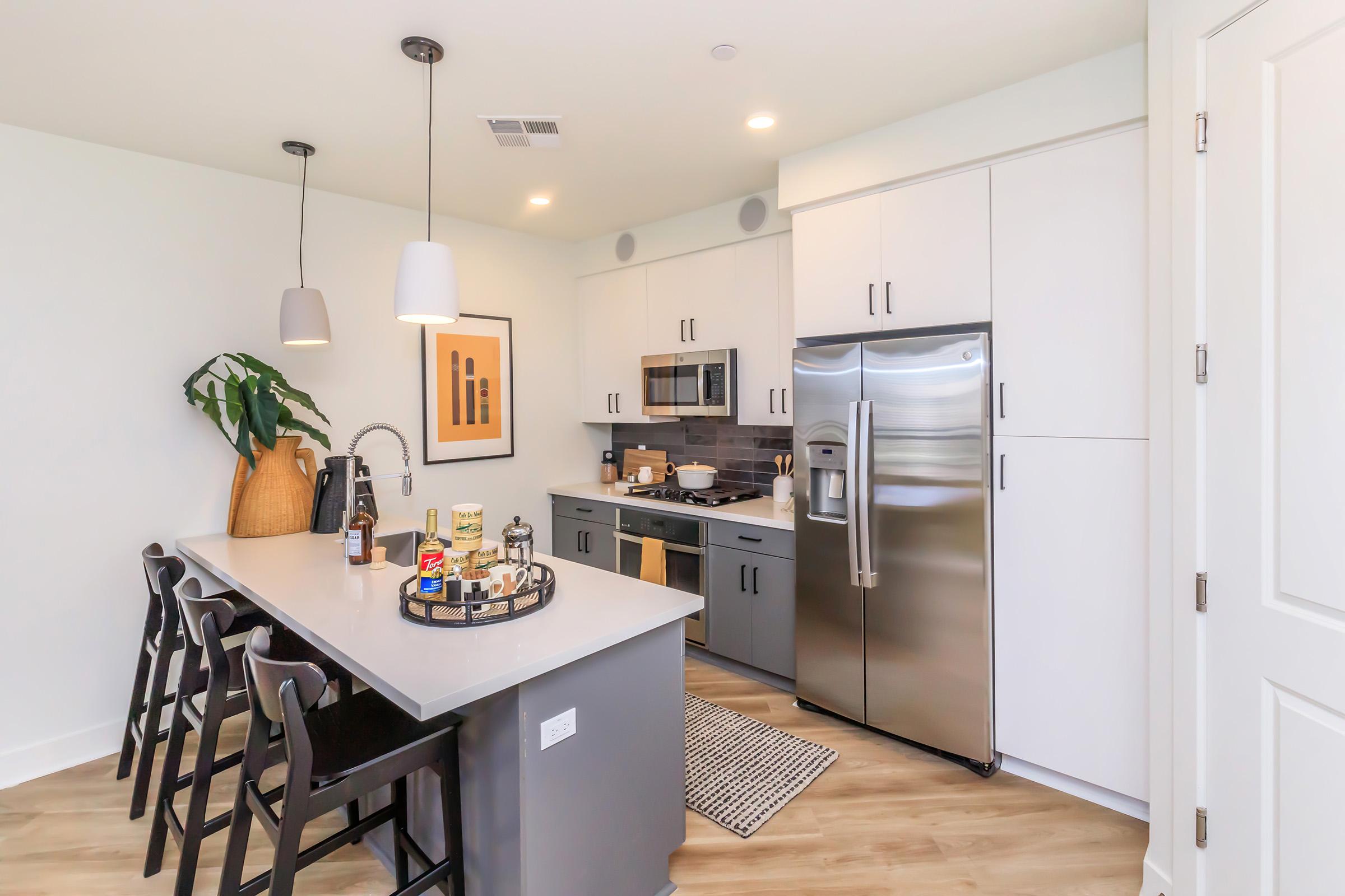 A modern kitchen featuring a spacious island with bar stools, stainless steel appliances, and white cabinetry. The decor includes a potted plant, kitchenware on the counter, and framed artwork on the wall, enhancing the contemporary aesthetic. The flooring is light wood, and the lighting is bright and inviting.