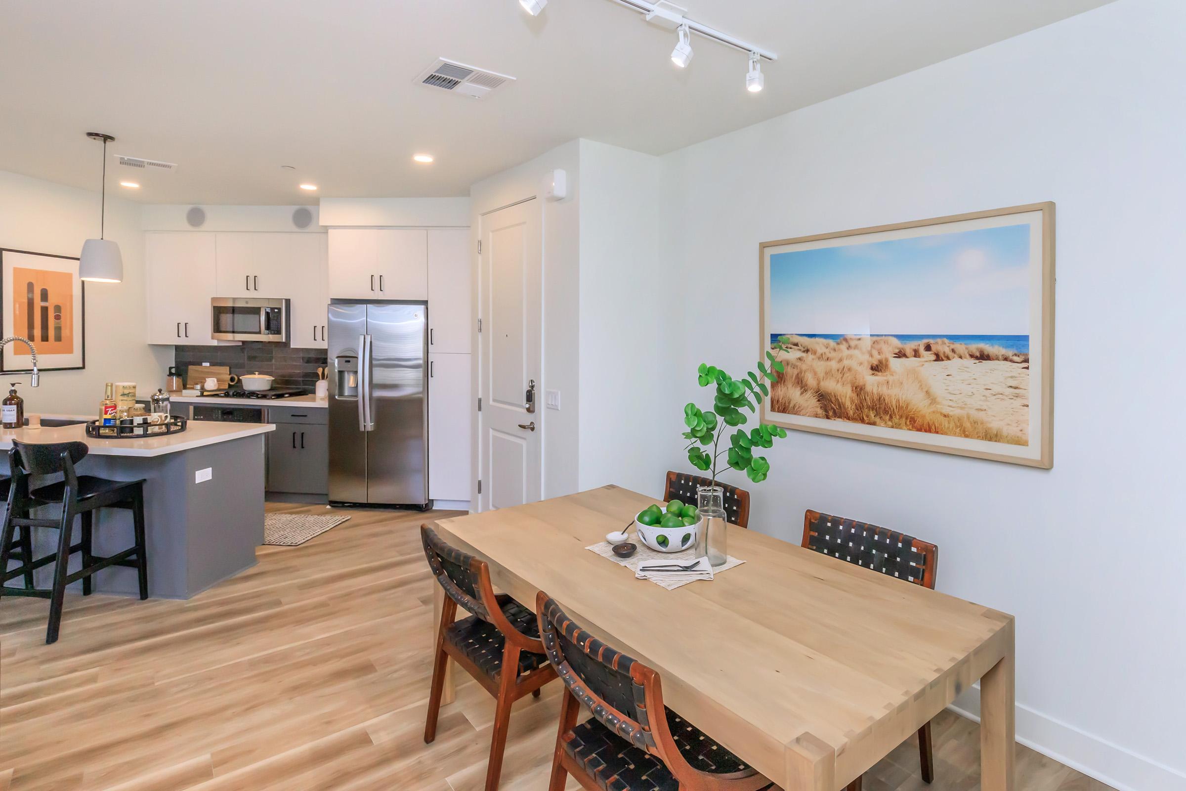 A modern kitchen and dining area featuring a wooden dining table with chairs, a contemporary kitchen with stainless steel appliances, and a large framed coastal landscape photograph on the wall. The space has light-colored walls and hardwood flooring, creating a bright and inviting atmosphere.