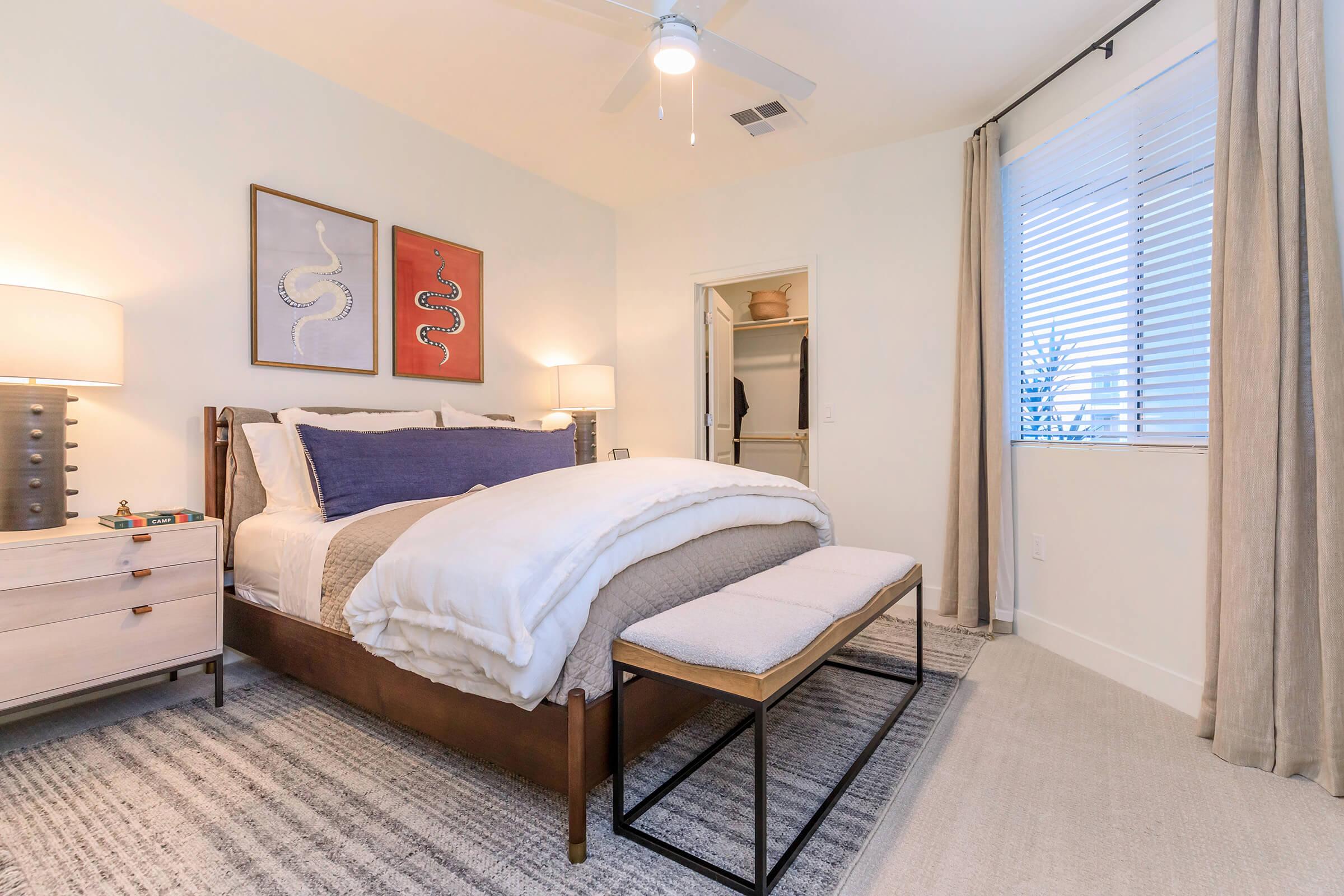 A modern bedroom featuring a wooden bed with white bedding and a blue accent pillow. Two snake-themed framed artworks hang above the bed. A nightstand with a lamp is on one side, and a bench is positioned at the foot of the bed. Light curtains frame a window with blinds, adding to the serene atmosphere.