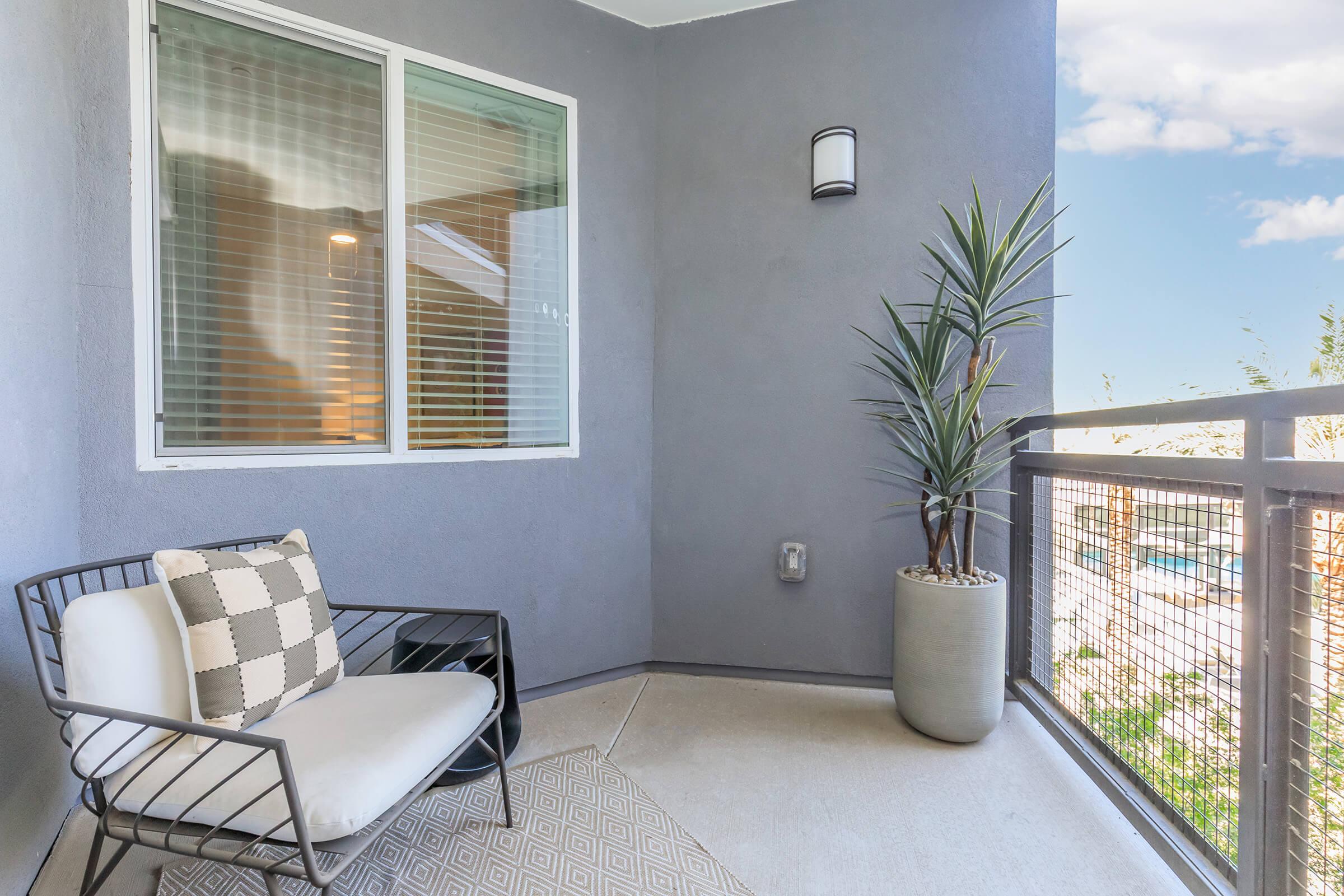 A modern balcony featuring a comfortable wire-frame chair with a cushion, a decorative potted plant, and a neutral-colored rug. The wall is painted in light gray, and there is a window with blinds in the background, allowing natural light to enter the space.