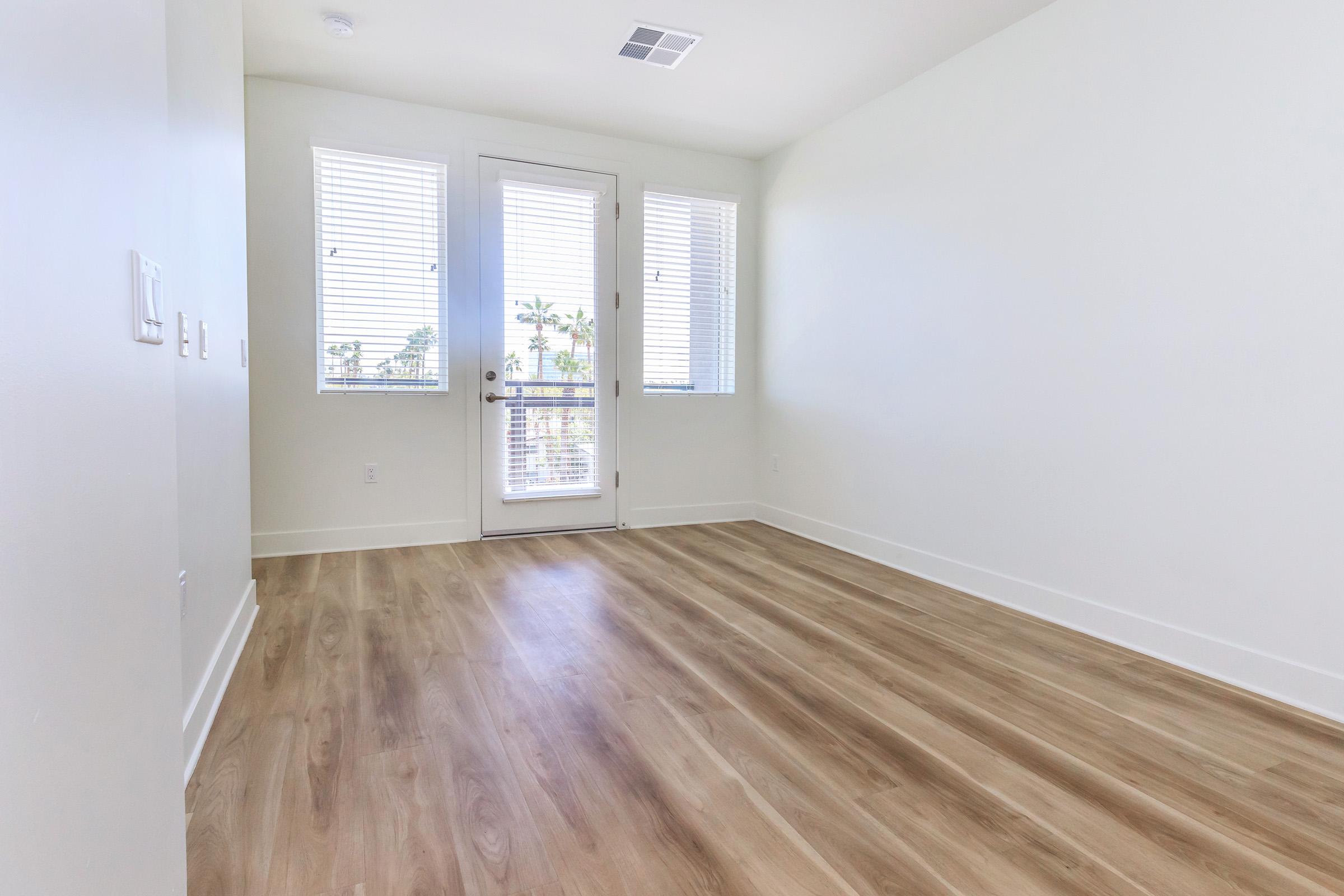 A spacious, empty room with light-colored walls and large windows that let in natural light. The flooring is a light wood laminate, and there is a glass door leading to a balcony or outdoor area. The overall atmosphere is bright and airy, ideal for personalizing or decorating.