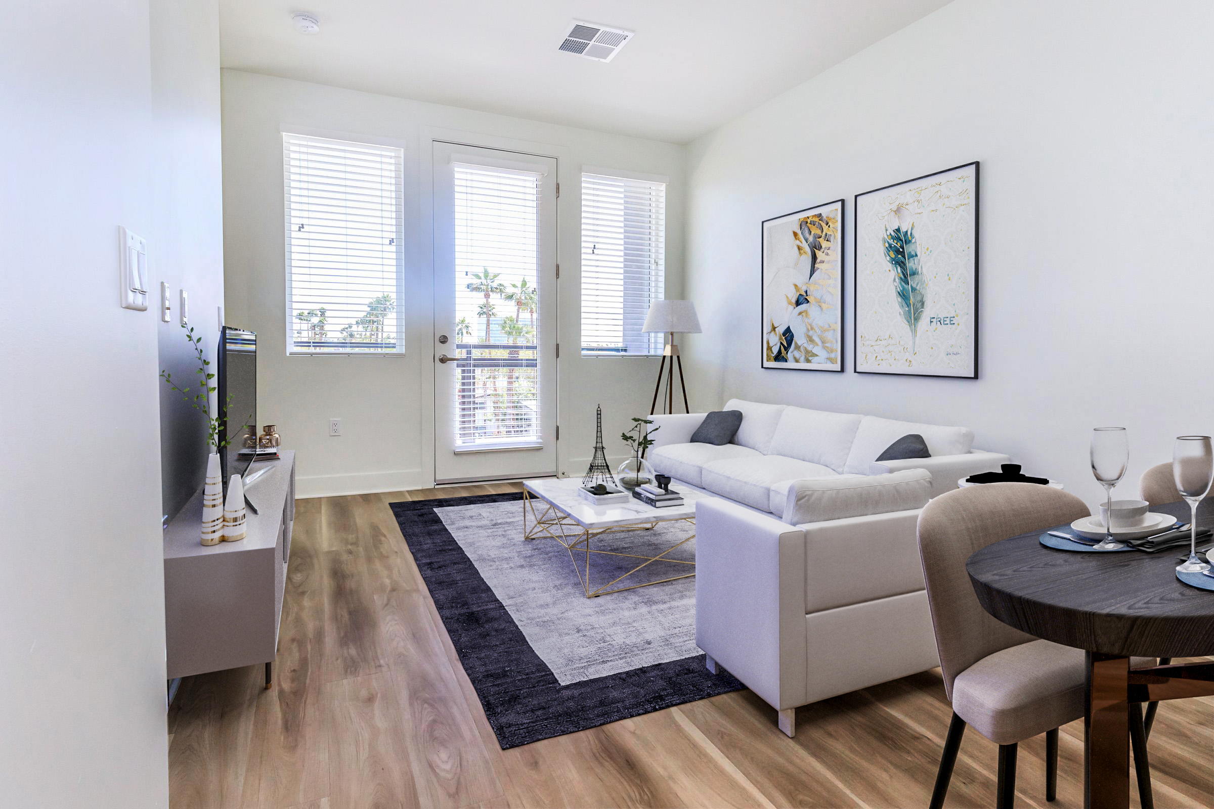 A bright and modern living room featuring a white sofa, a coffee table with decorative items, and a flat-screen TV. Large windows provide natural light, and two framed art pieces adorn the walls. A small dining table is set for two, enhancing the contemporary decor with stylish design elements.