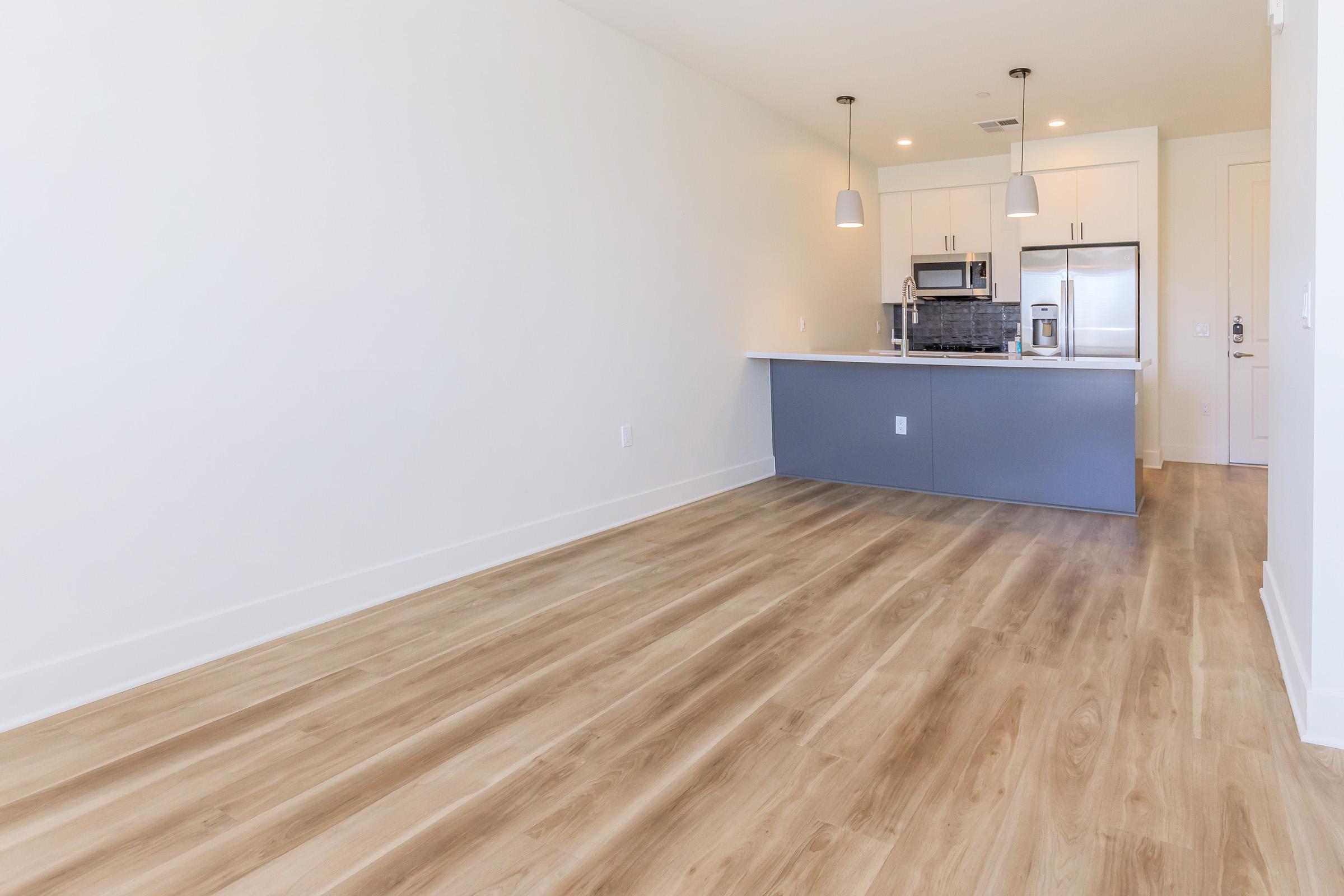 A modern, empty living space featuring light wooden flooring and white walls. In the background, a kitchen area with gray cabinetry, stainless steel appliances, and two pendant lights hanging above the countertop. The overall aesthetic is minimalistic and bright.