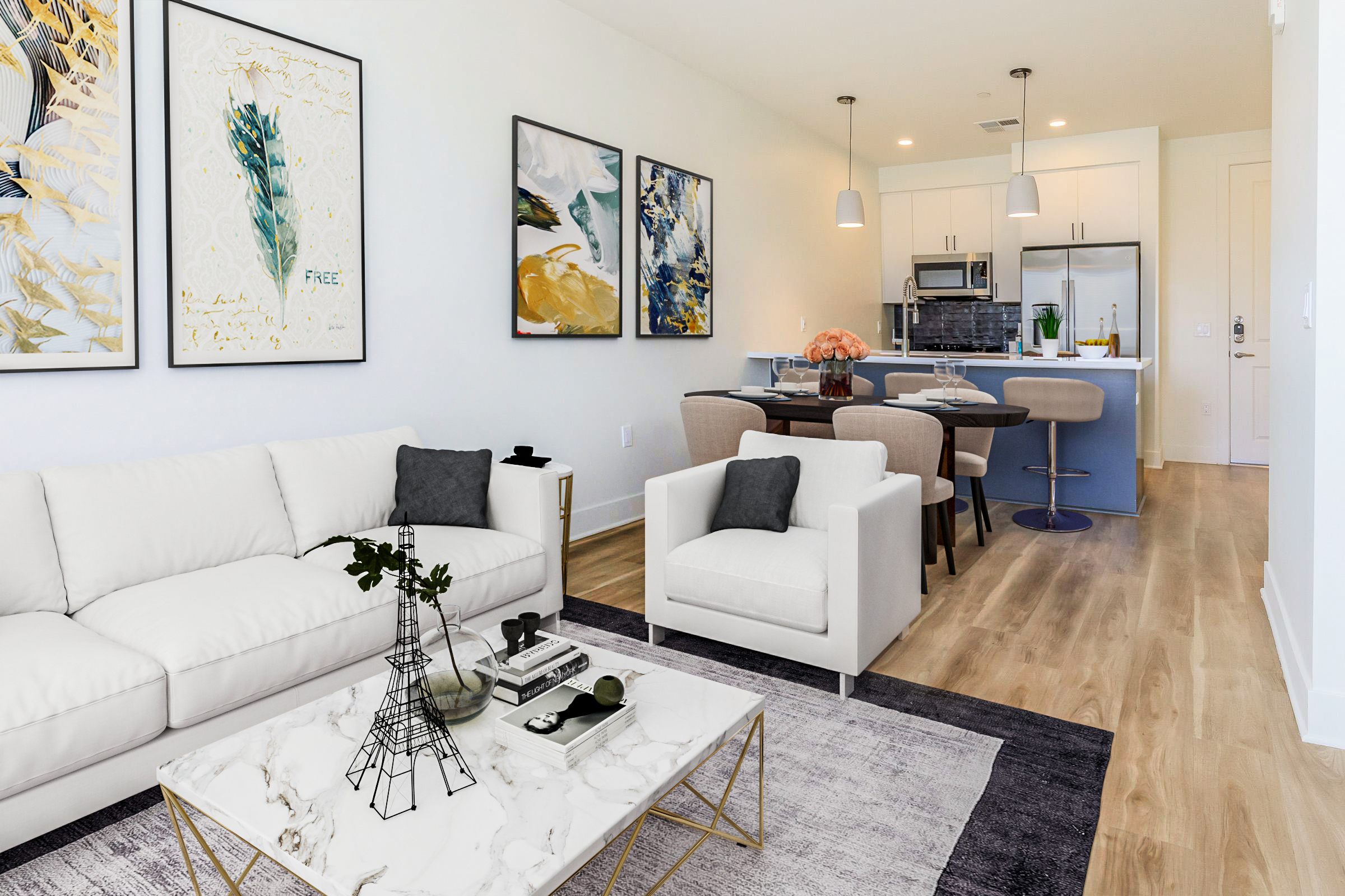 A modern living room featuring a white sofa and a stylish coffee table with decorative items. Artwork is displayed on the wall, and a dining area with a modern table and chairs is visible in the background. The space has wooden flooring and a bright, airy ambiance.