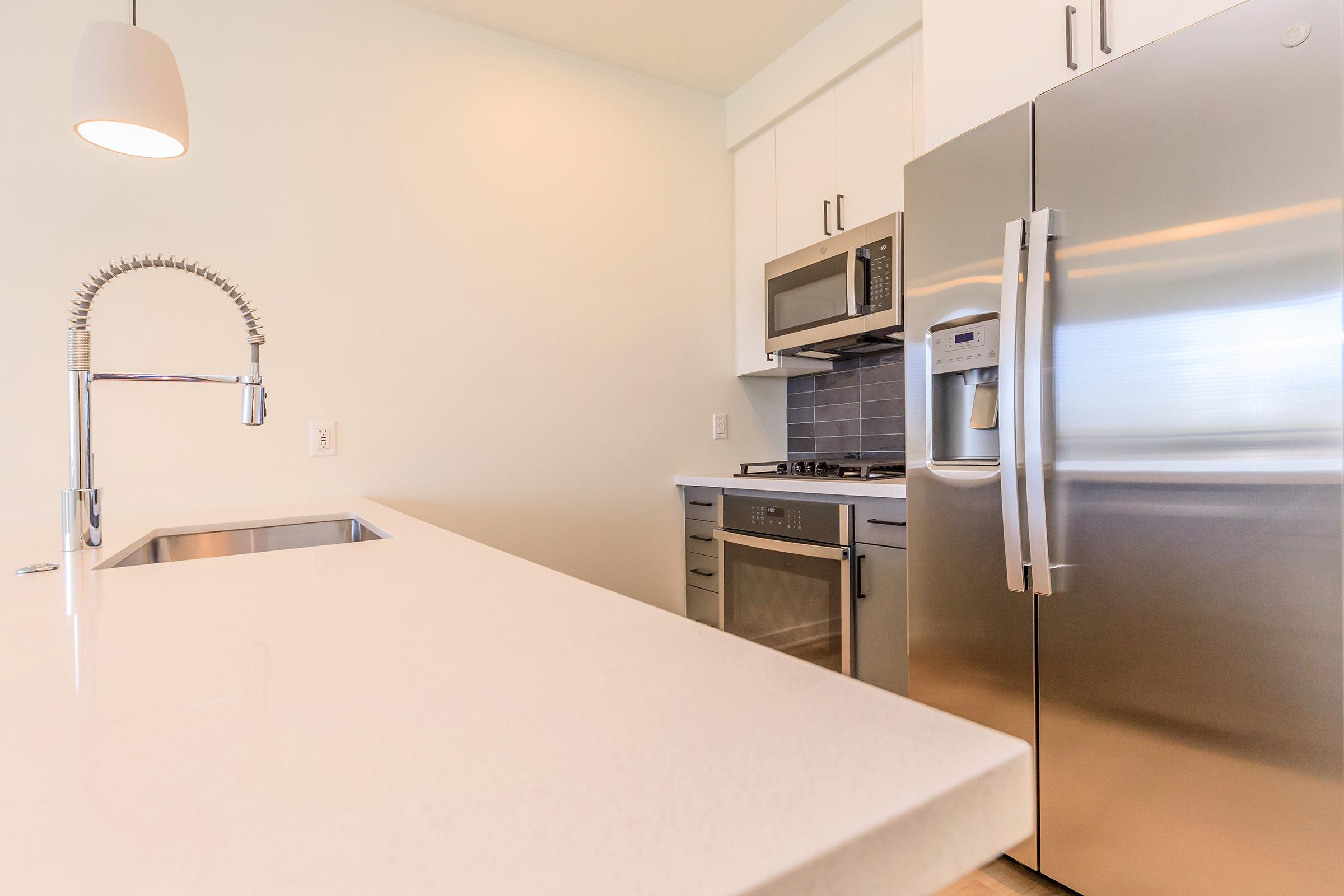 A modern kitchen featuring a sleek countertop and stainless steel appliances, including a refrigerator, microwave, and oven. A contemporary sink with a pull-out faucet is visible in the foreground, and there is a pendant light hanging from the ceiling, providing a bright and open atmosphere.