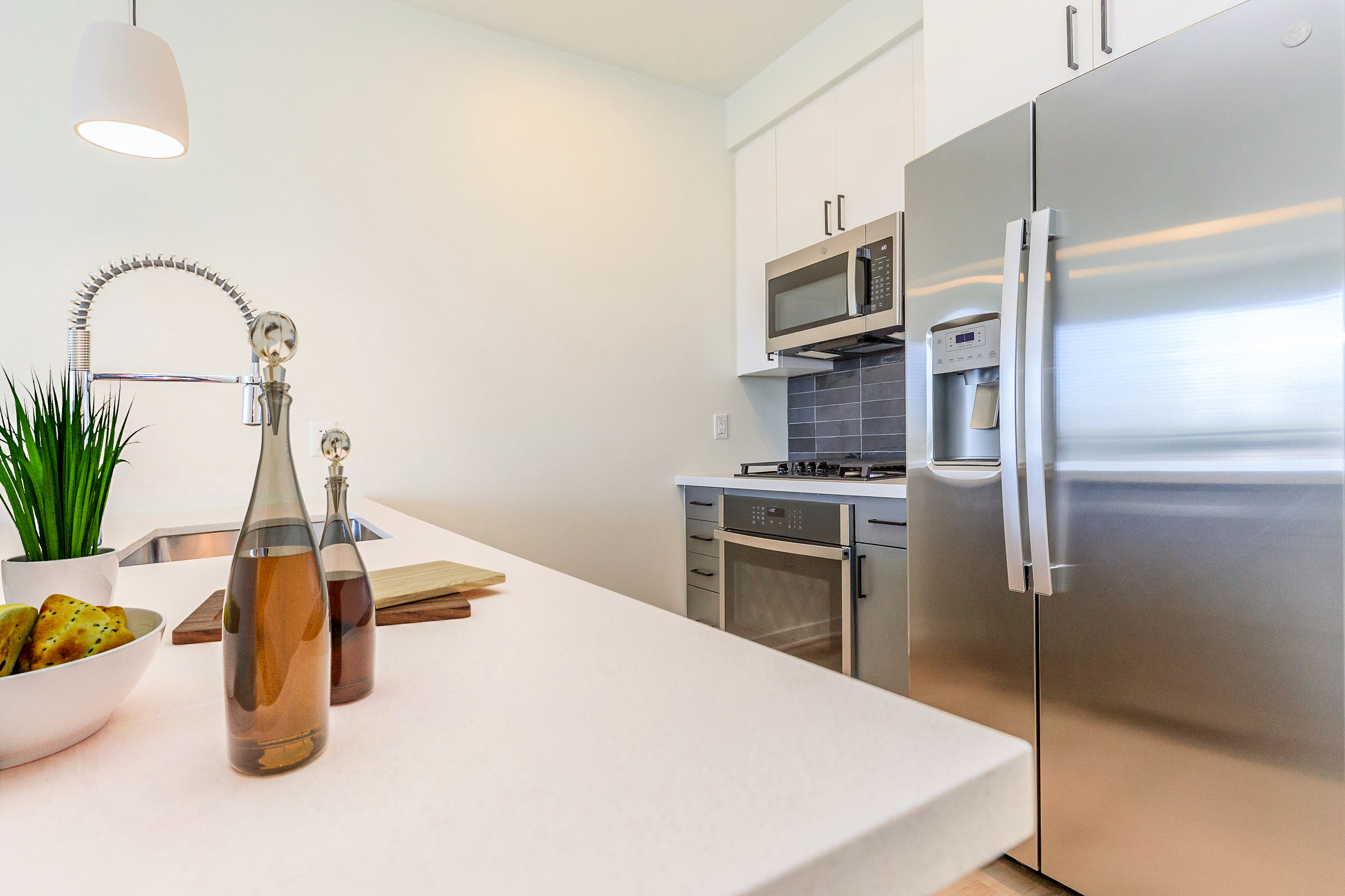 Modern kitchen featuring a sleek countertop with two glass bottles and a bowl of green apples. A stainless steel refrigerator and microwave are visible, along with a sink with a stylish faucet. Minimalistic design with light colors and a touch of greenery in the background.