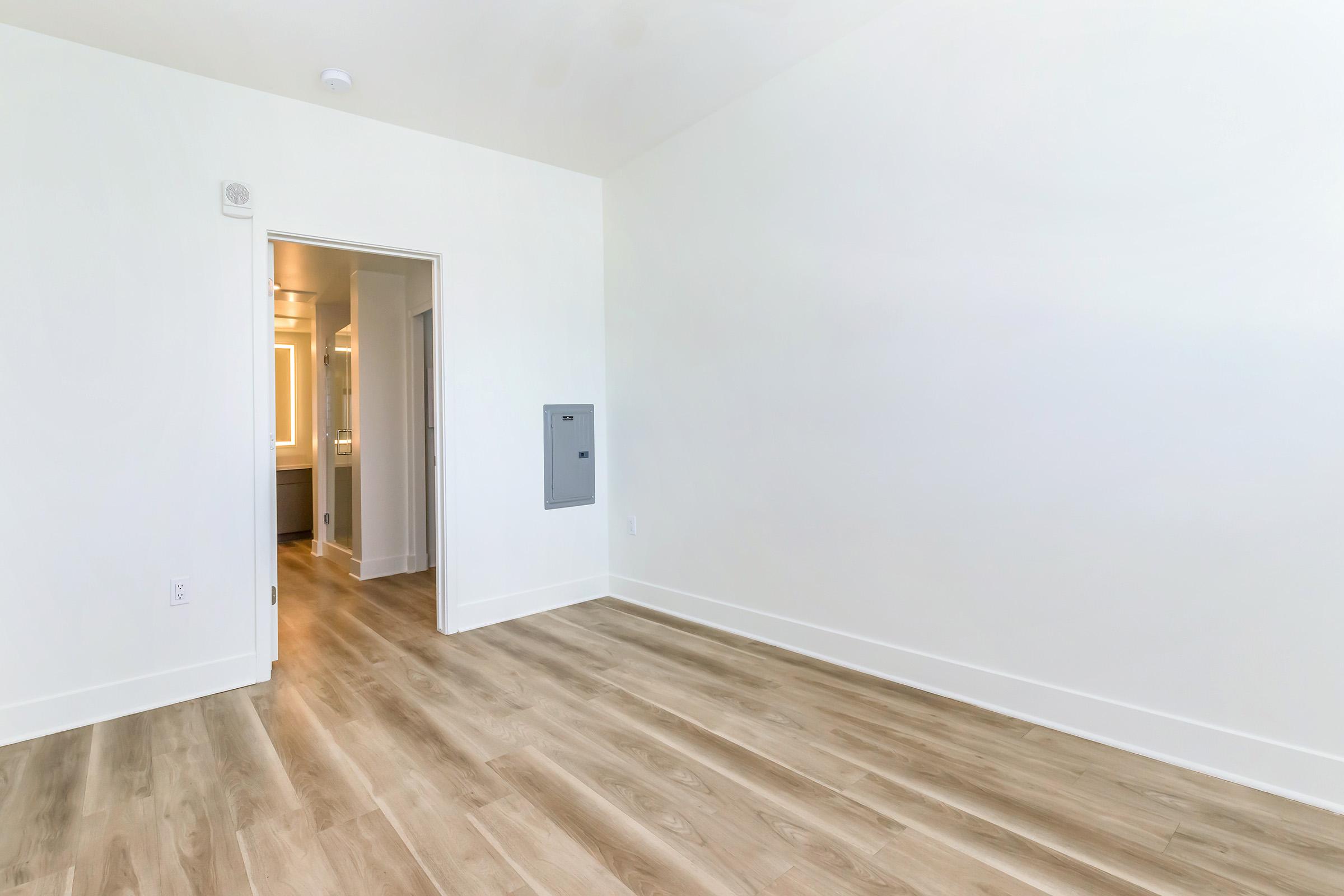 A minimalistic interior of a room featuring light-colored walls and a wooden floor. An open doorway leads to another space, possibly a bathroom or hallway, with soft lighting visible. There is a mail slot or utility panel on the wall. The overall ambiance is bright and spacious.