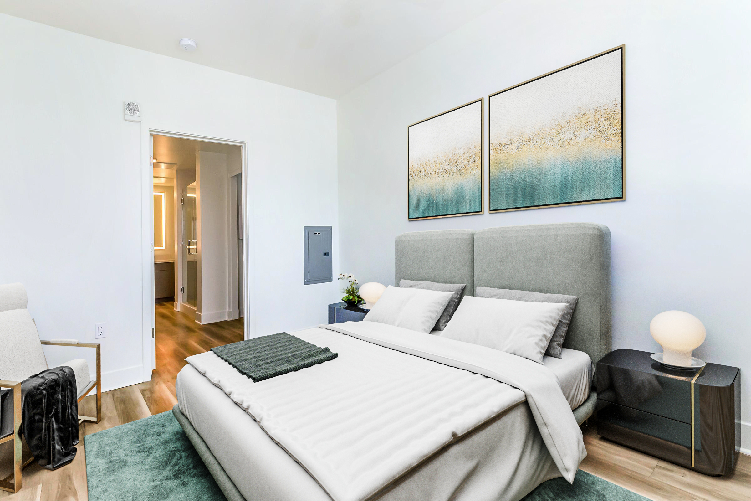 A modern bedroom featuring a neatly made bed with light gray bedding and decorative pillows. There are two abstract paintings above the bed, a nightstand with a soft-glow lamp, and a plant. A chair sits in the corner, and there's an open door leading to a bathroom area visible in the background. The walls are light and the flooring is wooden.