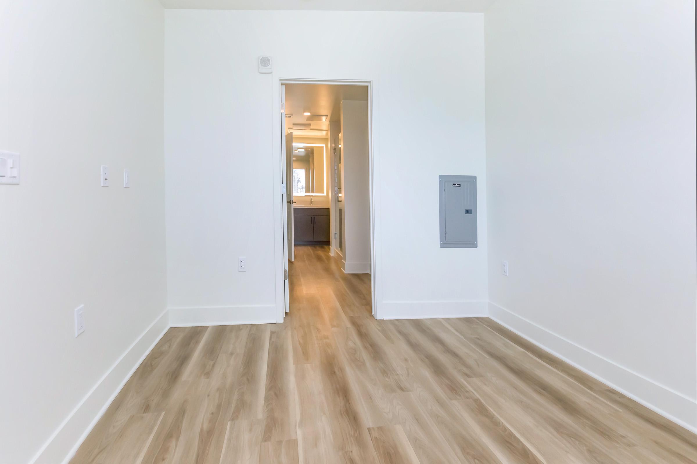 A spacious, empty room with light-colored walls and wooden flooring. A doorway leads to a bathroom area in the background, and there is an electrical panel on the right wall. The room features natural light streaming in, creating a bright and airy atmosphere.
