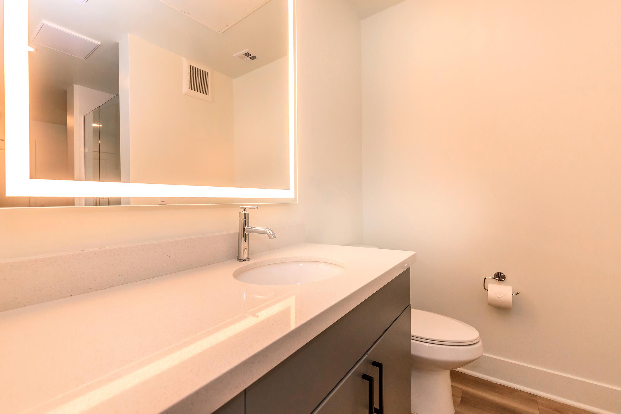 Modern bathroom featuring a sleek countertop with a circular sink, a large illuminated mirror above, and minimalistic decor. There is a toilet in the corner and a towel holder. The walls are light-colored and the flooring is wood-like.