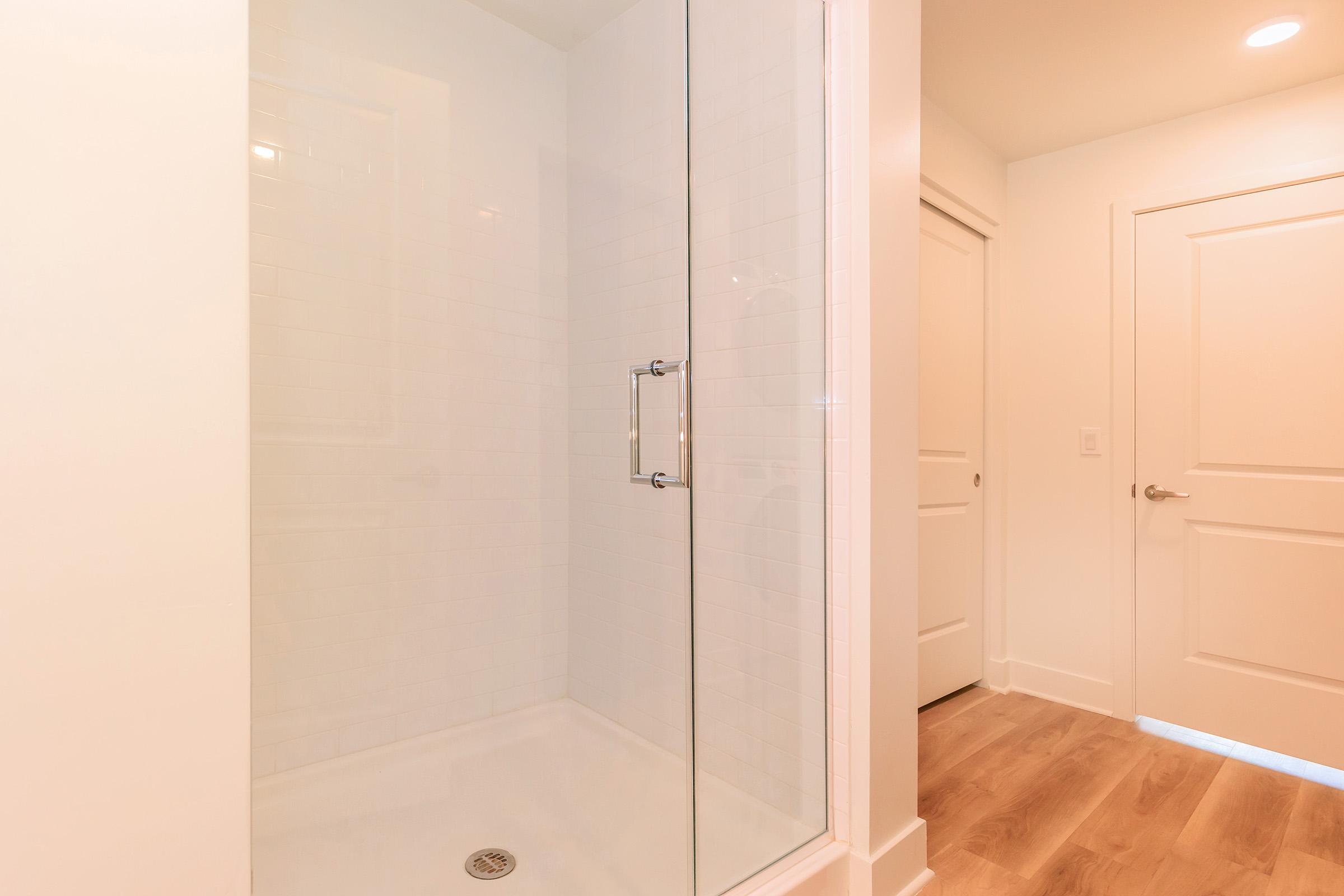 A clean and modern shower with a glass door, featuring white tiled walls and a tile floor. The scene includes a light-colored hallway with a closed door on the right and a light fixture above, showcasing a bright and minimalistic bathroom design.