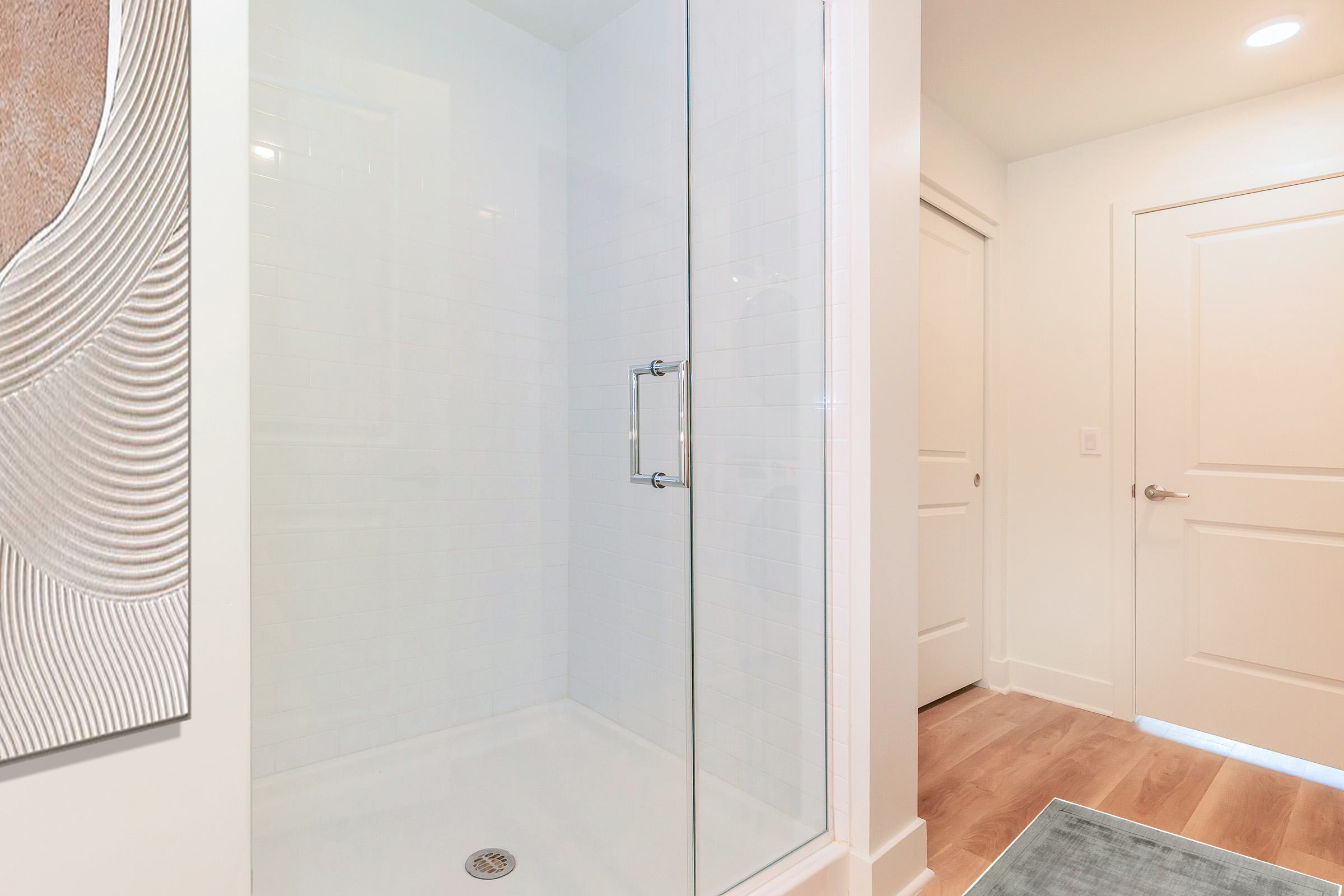 A modern bathroom featuring a clear glass shower enclosure on the left, with white tiled walls and a textured artwork on the adjacent wall. On the right, there's a white door leading to another room and a light-colored floor. The overall aesthetic is clean and contemporary.