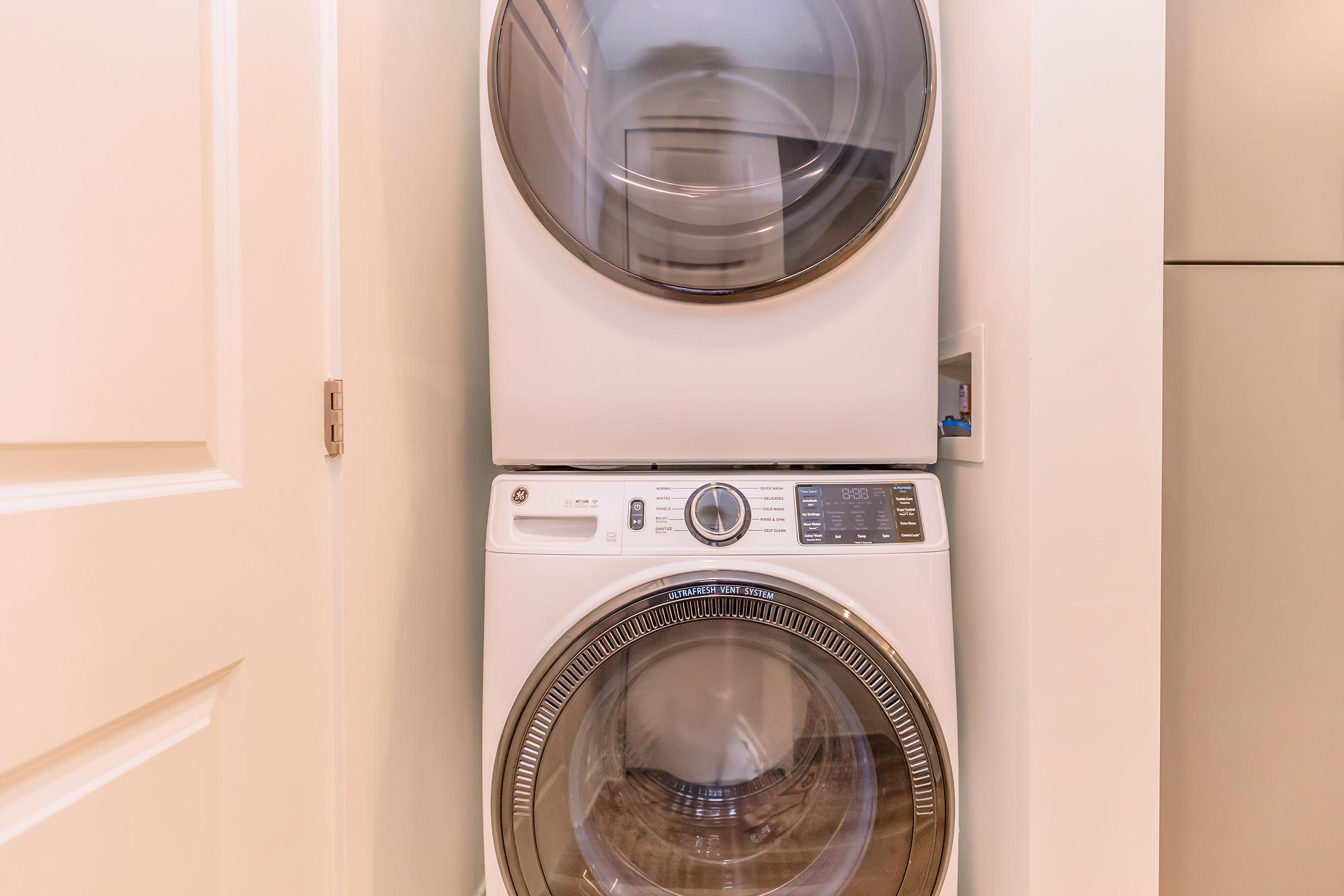 A stacked laundry unit featuring a front-loading washer on the bottom and a dryer on top, both with a sleek white finish. The appliances are situated in a well-lit laundry area, with a cabinet door visible in the background.
