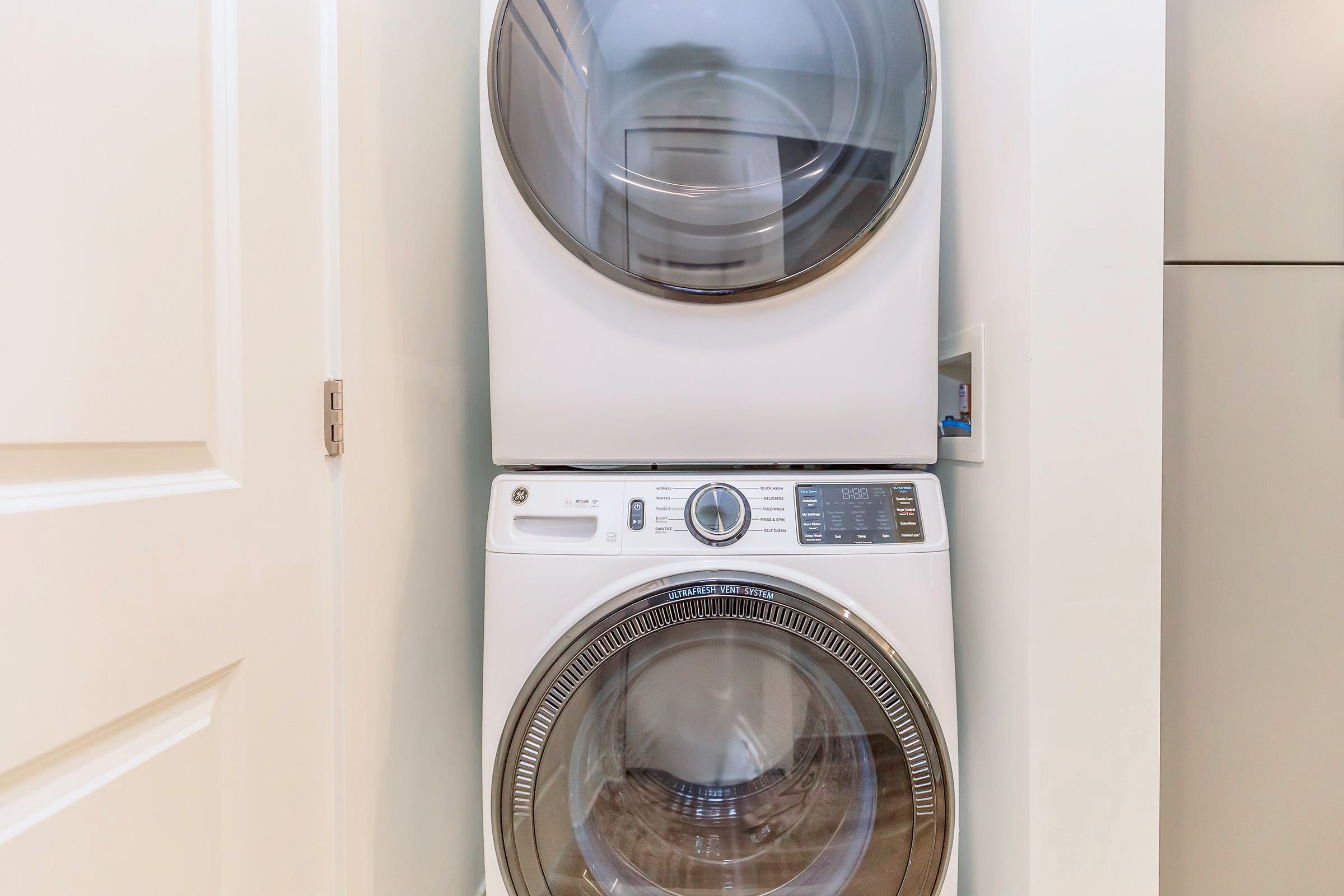 A stacked washer and dryer set in a laundry nook. The appliances are white with modern designs, featuring digital displays and large glass doors. The surrounding walls are light-colored, complementing the sleek look of the machines. The space appears clean and organized.