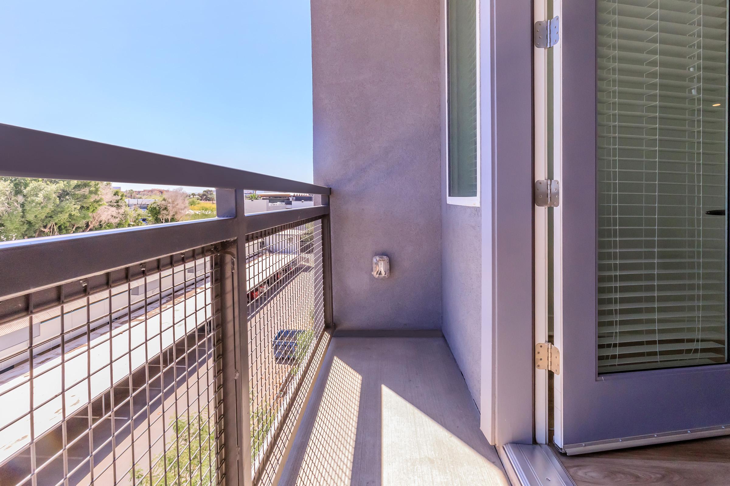 A corner of a modern balcony with a metal railing, showing a clear blue sky and distant greenery. The balcony features a concrete floor and a glass door that leads indoors, providing a bright, open feel. Sunlight casts soft shadows on the balcony surface.