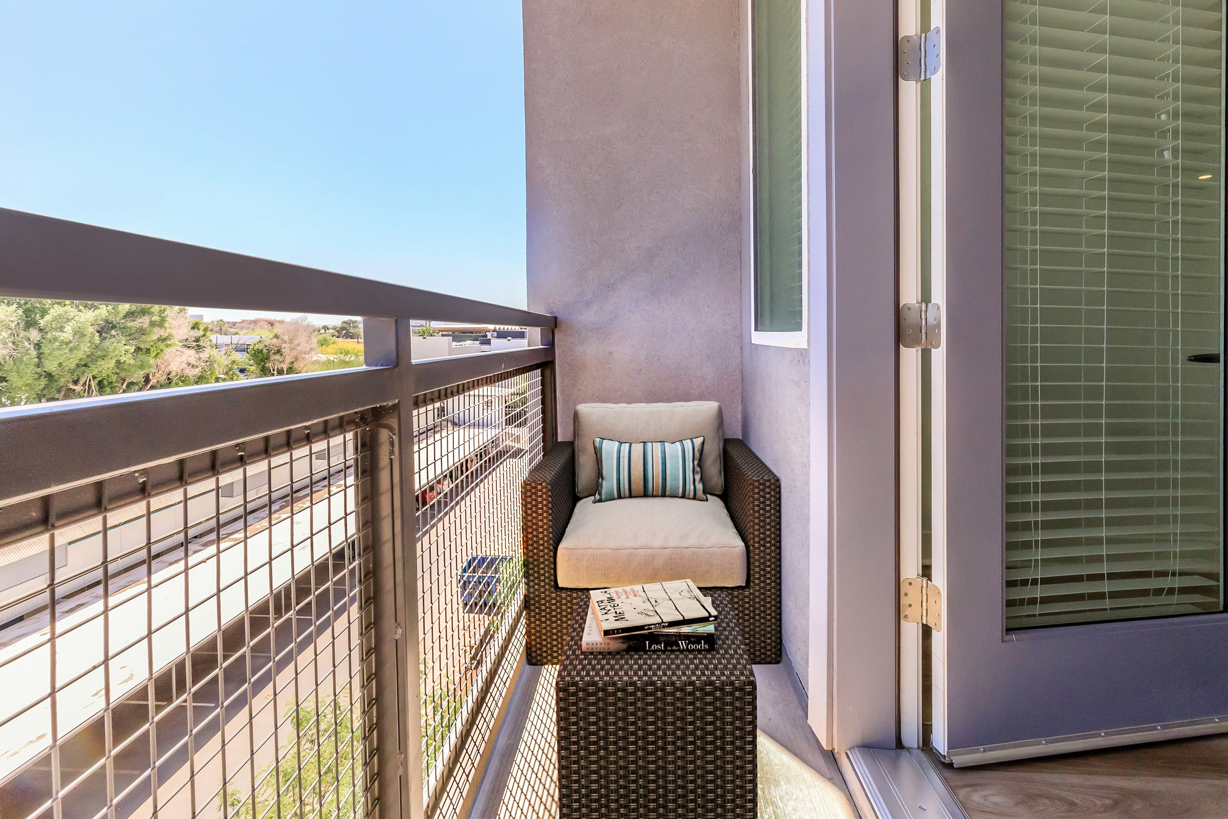 A small balcony features a comfortable chair with a light-colored cushion and decorative striped pillow, positioned next to a square table holding a stack of magazines. The railing has a modern design, and the view includes trees and a clear blue sky. Natural light enhances the inviting atmosphere.