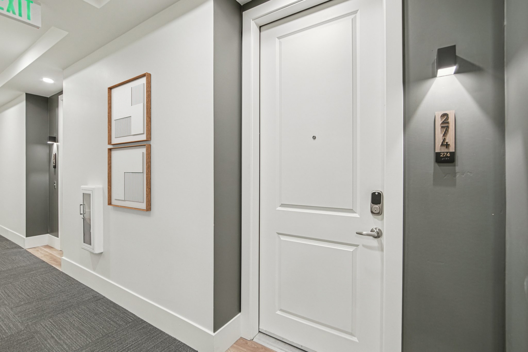 A hallway featuring a white door labeled "274" with a modern door handle, next to a wall-mounted light. Two framed artworks hang on the adjacent wall, complementing the neutral color scheme and carpeted floor, creating a contemporary living space ambiance.
