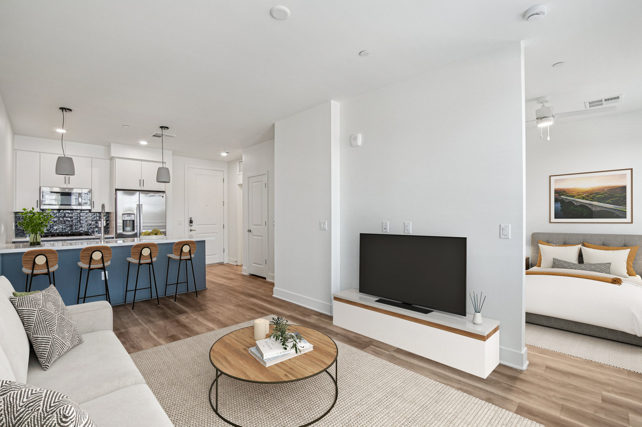 A modern apartment interior featuring a cozy living area with a light-colored sofa, a round coffee table, and a large flat-screen TV. The kitchen area has bar stools and sleek cabinets. A well-lit space with neutral colors and a small plant adds a touch of warmth. A bedroom is visible in the background, separated by a partial wall.