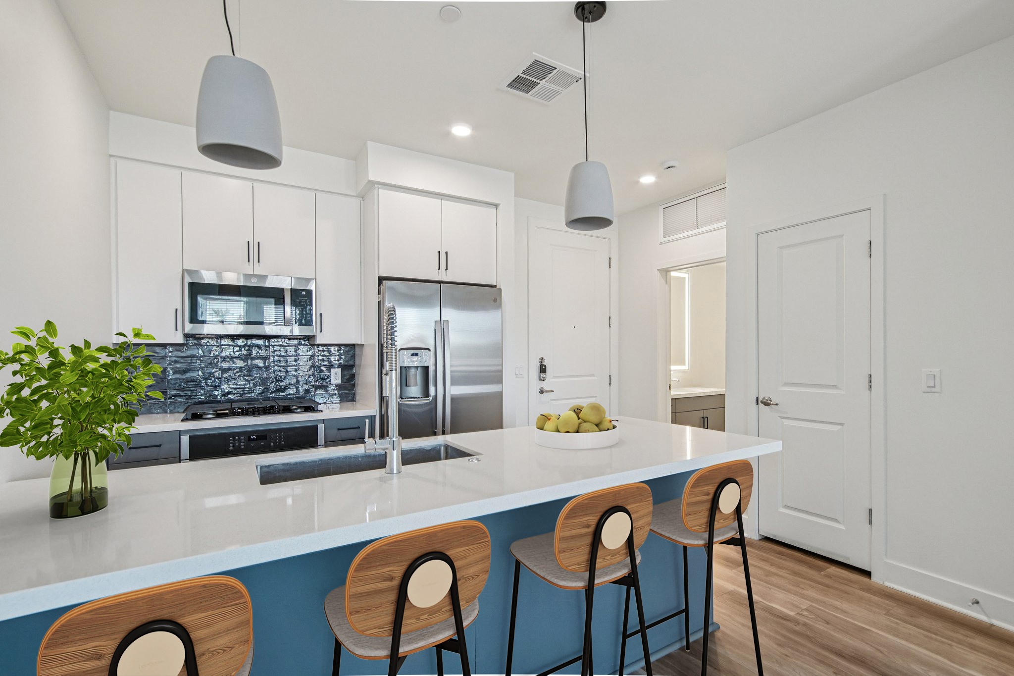 Modern kitchen featuring a large island with four wooden stools, sleek stainless steel appliances, and white cabinetry. A bowl of green apples sits on the countertop, complemented by pendant lighting. The room has a bright, airy feel with natural light coming from a nearby window.