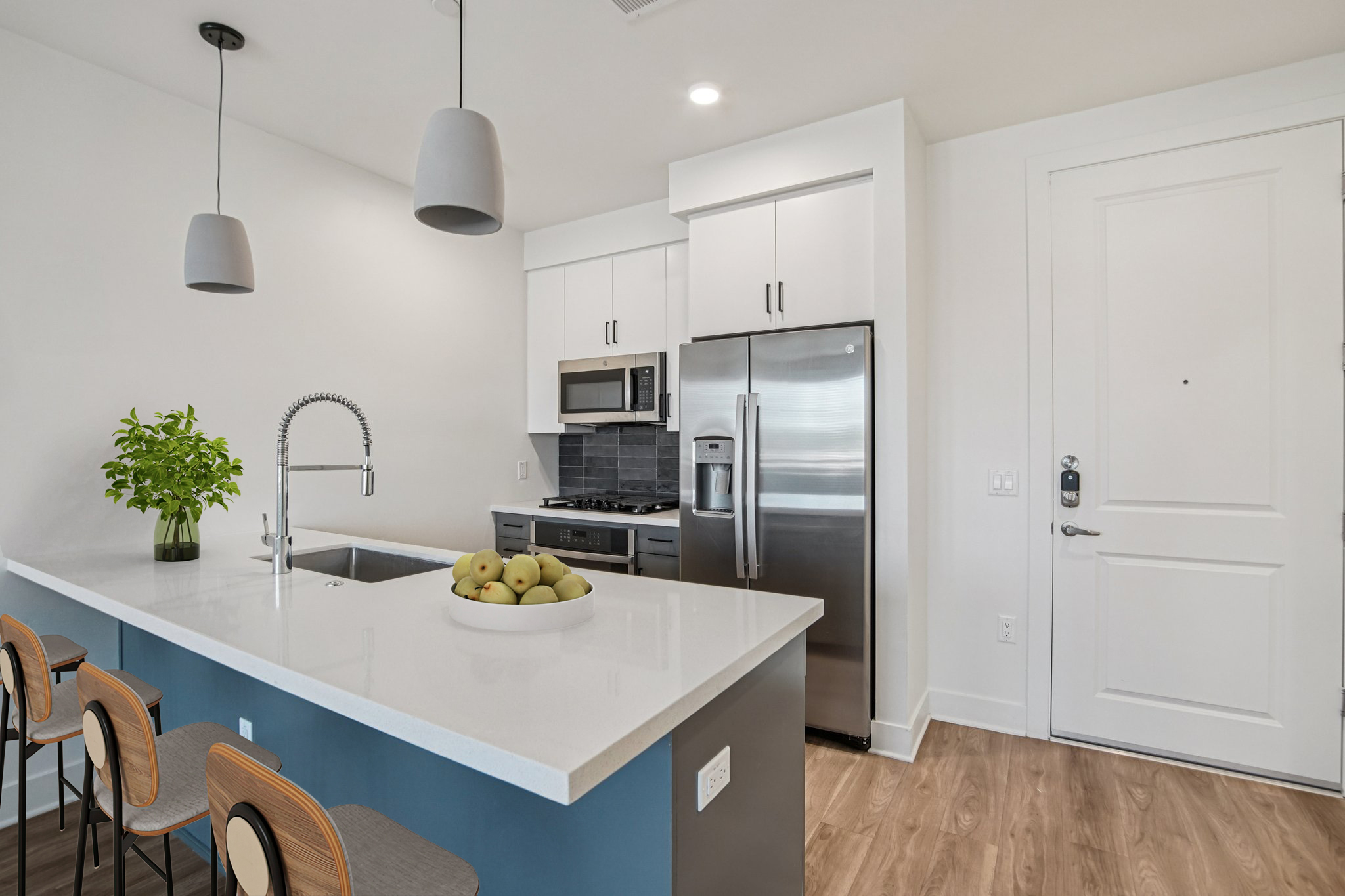 Modern kitchen with a central island featuring a bowl of green apples, bar stools, stainless steel appliances including a refrigerator and microwave, and minimalist cabinetry. The space is well-lit with pendant lamps and has a contemporary design, showcasing light wood flooring and a sleek entrance door.