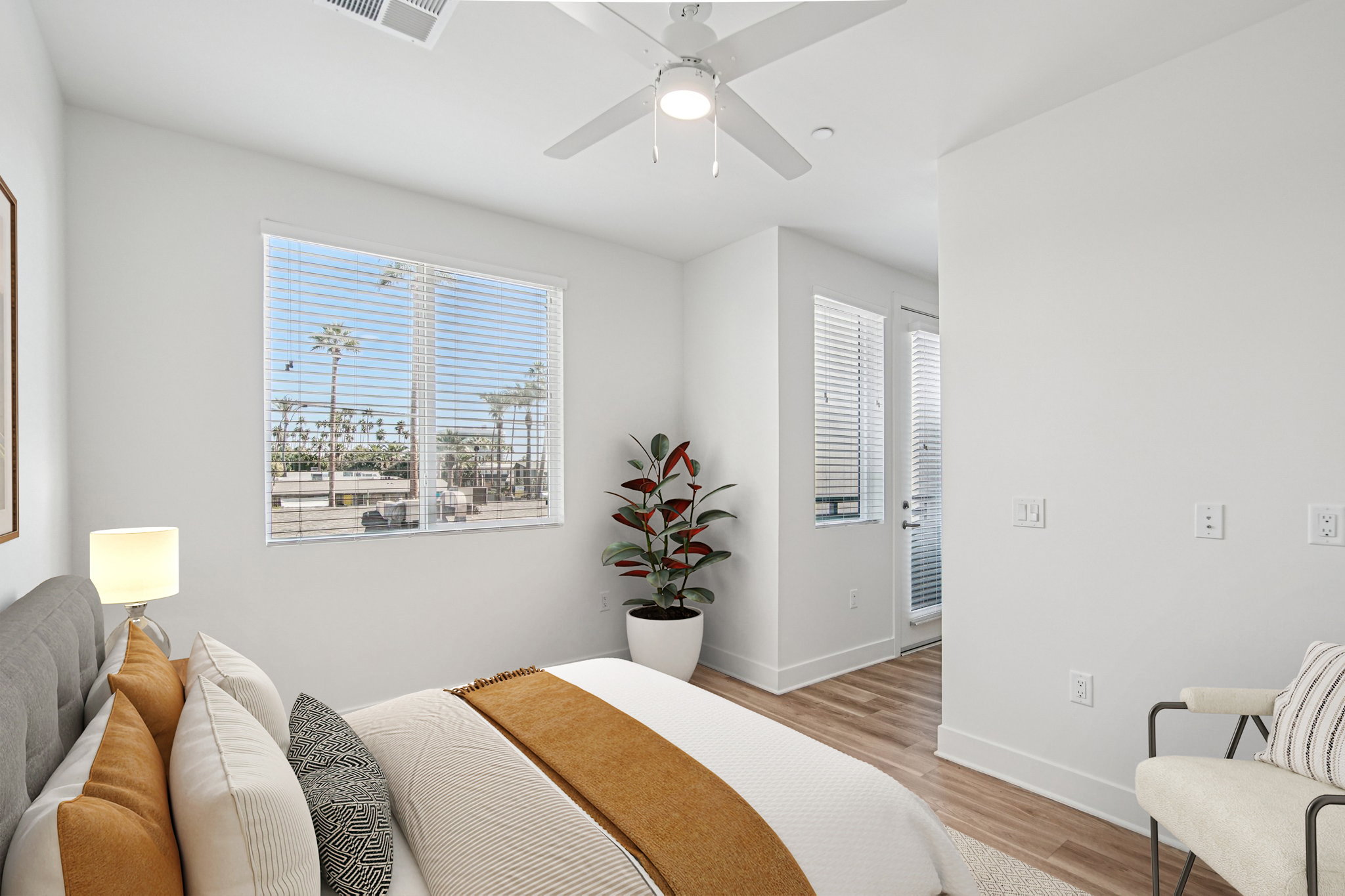A bright and airy bedroom featuring a neatly made bed with a gray headboard and tan throw blanket. There’s a ceiling fan, large window with white blinds, and a potted plant in the corner. A door leads to an adjoining area, with light wood flooring throughout the space.