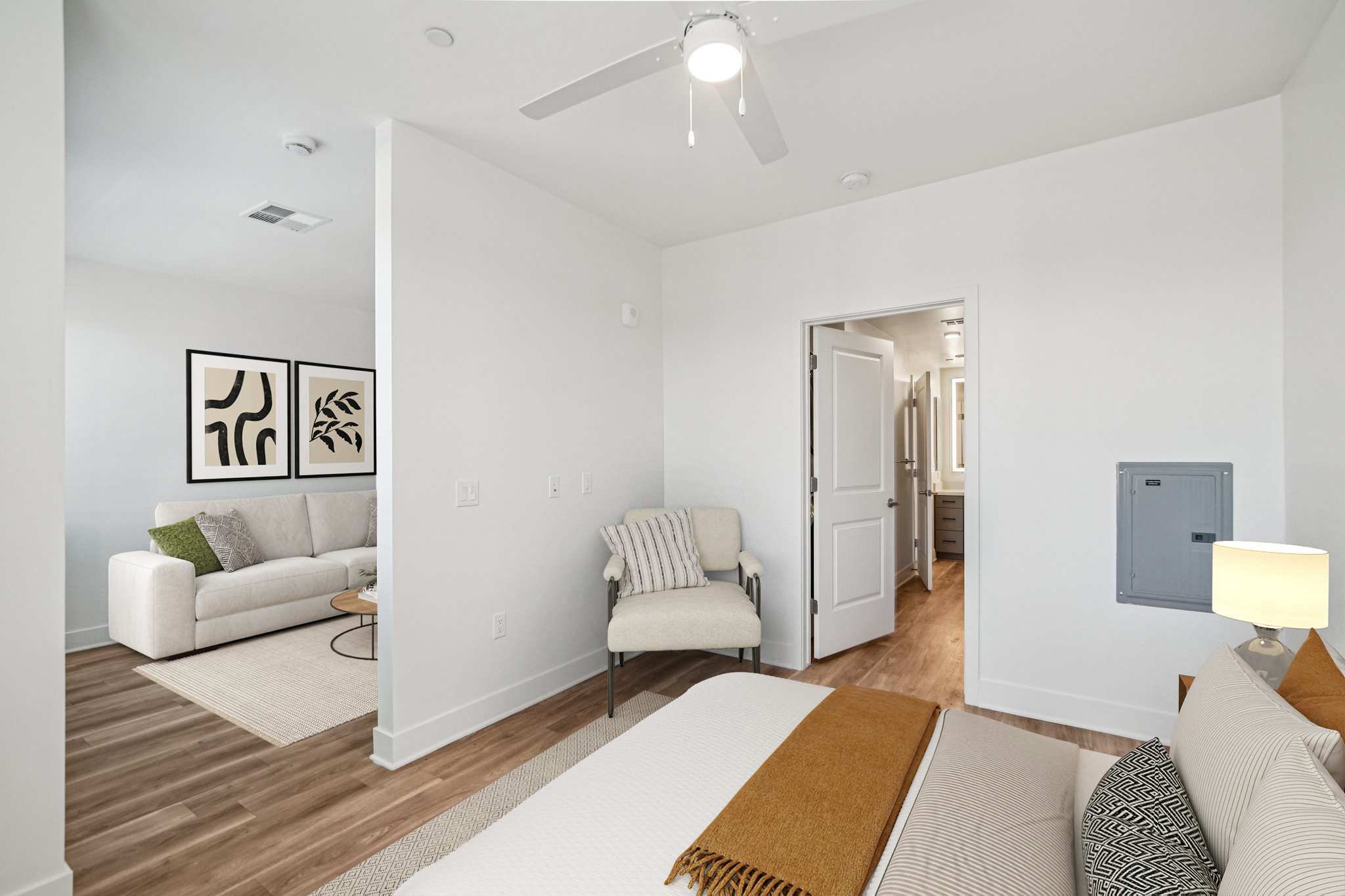 A modern bedroom featuring a bed with a light-colored blanket, a beige chair, and a small lamp on a nightstand. In the background, there's a living area with a white couch and framed artwork on the wall. The room has light-colored walls and wooden flooring, creating a bright and inviting atmosphere.