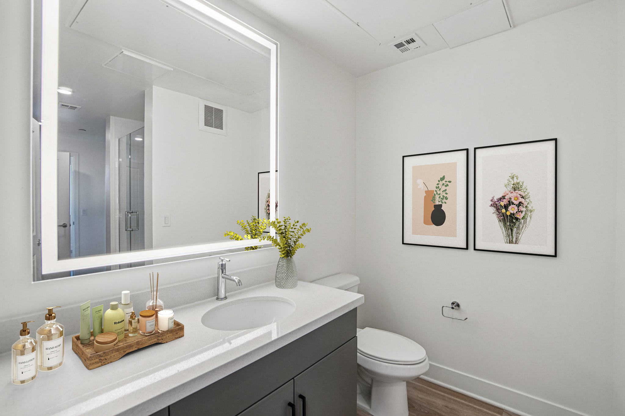 A modern bathroom featuring a sleek gray vanity with a circular sink. Above the sink, a large illuminated mirror reflects the space. Decorative bottles and a small tray with skincare products sit on the vanity. To the right, two framed art pieces depict minimalist floral designs. The walls are painted white, creating a clean aesthetic.
