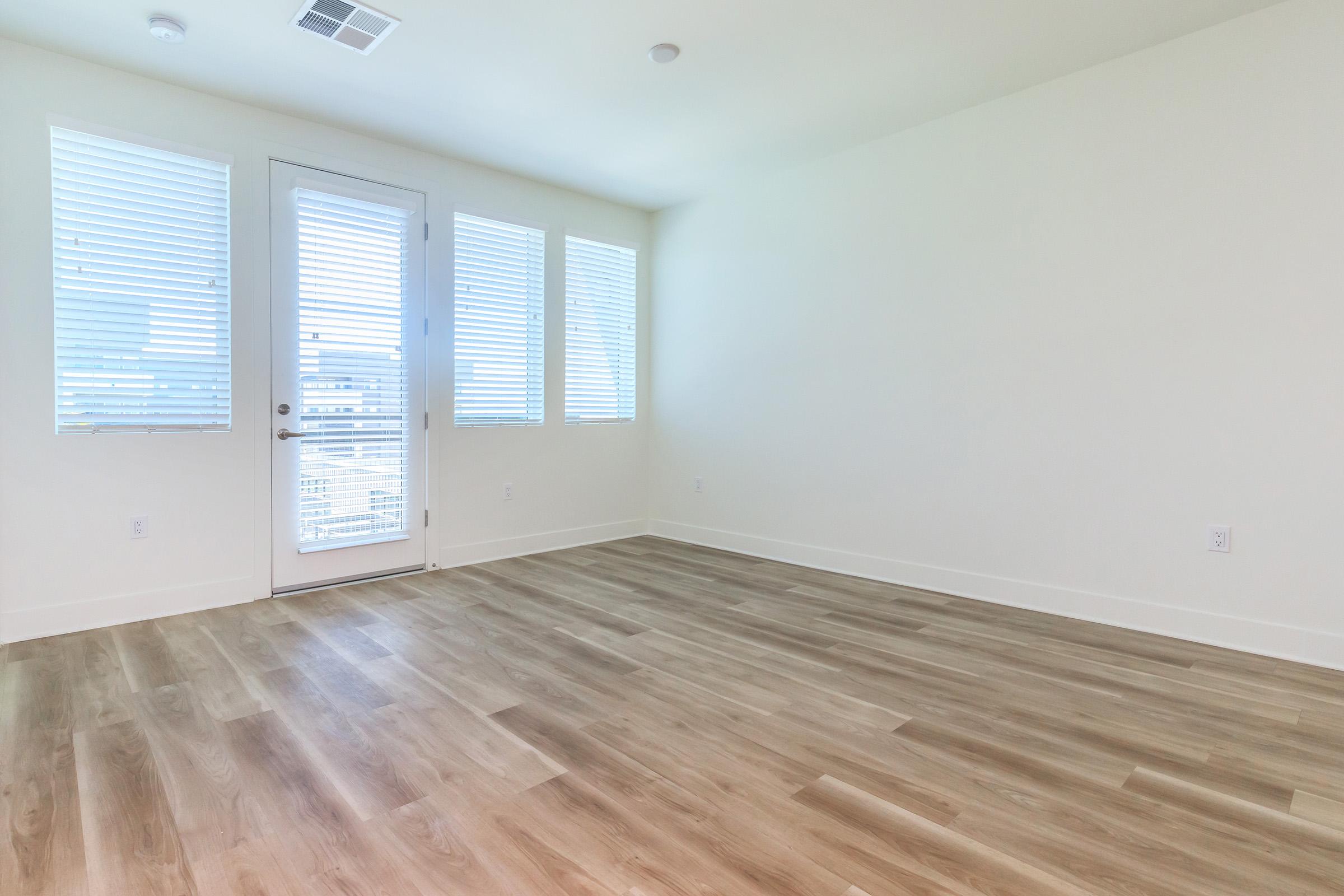 A bright, empty room with light-colored walls and wooden flooring. Large windows with blinds allow natural light to enter. A glass door leads to a balcony or outdoor area, creating an open, spacious feel. The room is ready for furnishing and personalization.