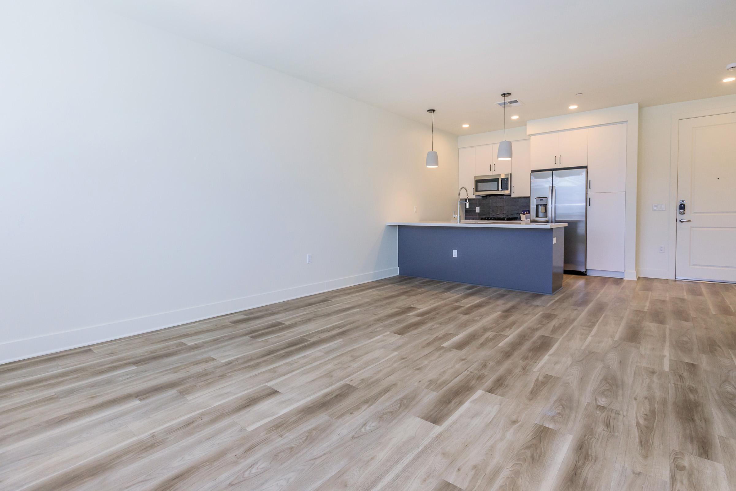 A spacious, modern kitchen area with light-colored walls and wooden flooring. The kitchen features a grey island, stainless steel appliances, and pendant lighting. The living space is open and bright, with a door leading to the outside visible in the background.