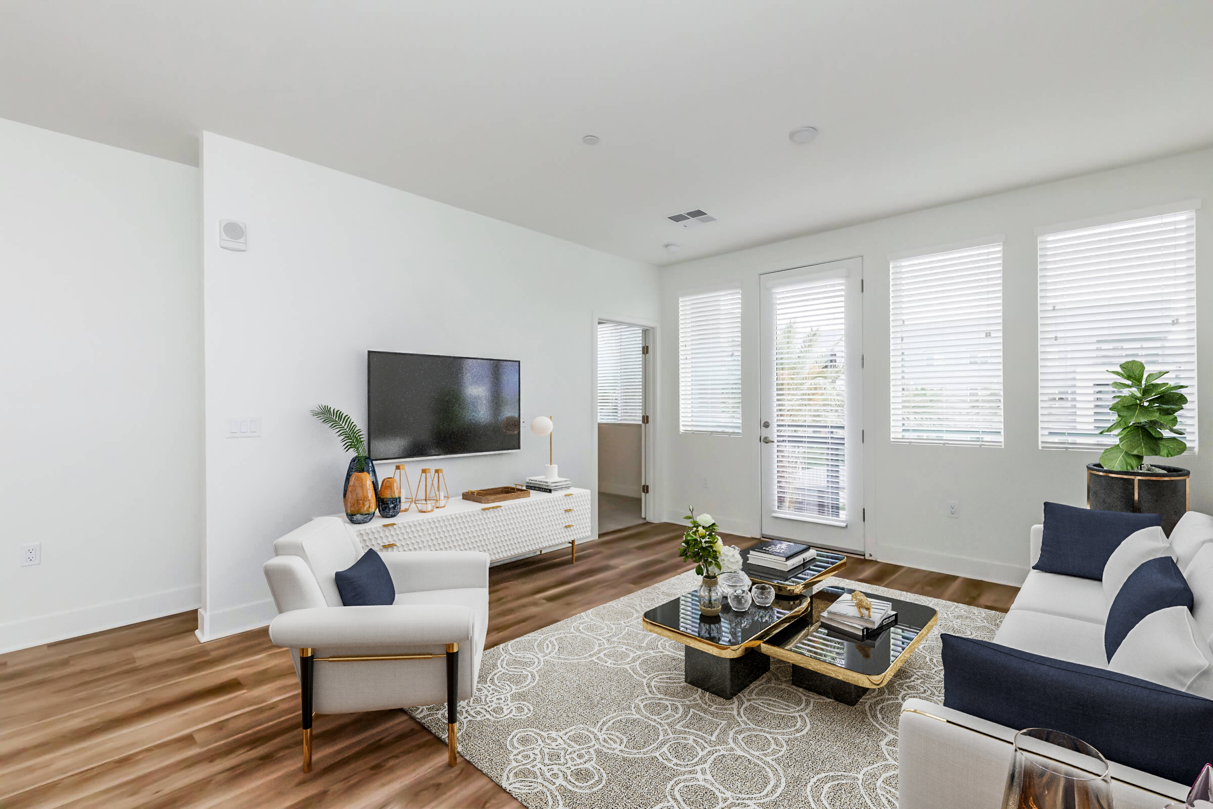 A modern living room featuring a light color palette. The space includes a comfortable sofa with blue and white cushions, a coffee table with books and decorative items, a television mounted on the wall, and large windows allowing natural light. A potted plant adds a touch of greenery.