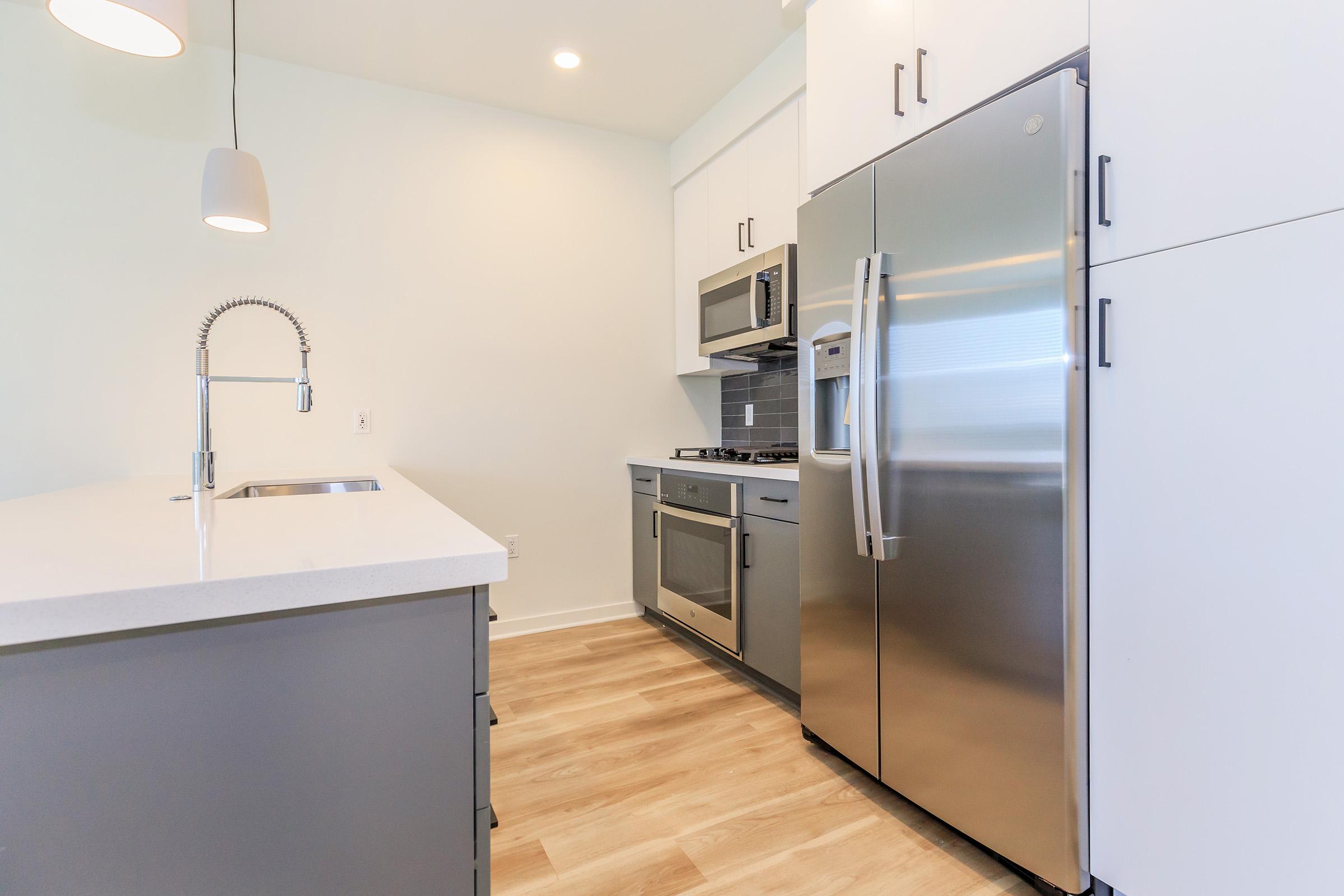 Modern kitchen featuring stainless steel appliances, including a refrigerator, microwave, and oven. The design includes white cabinets and a light-colored countertop with a sink. A pendant light hangs above the kitchen island, and the flooring is wood-like laminate. Natural light fills the space.