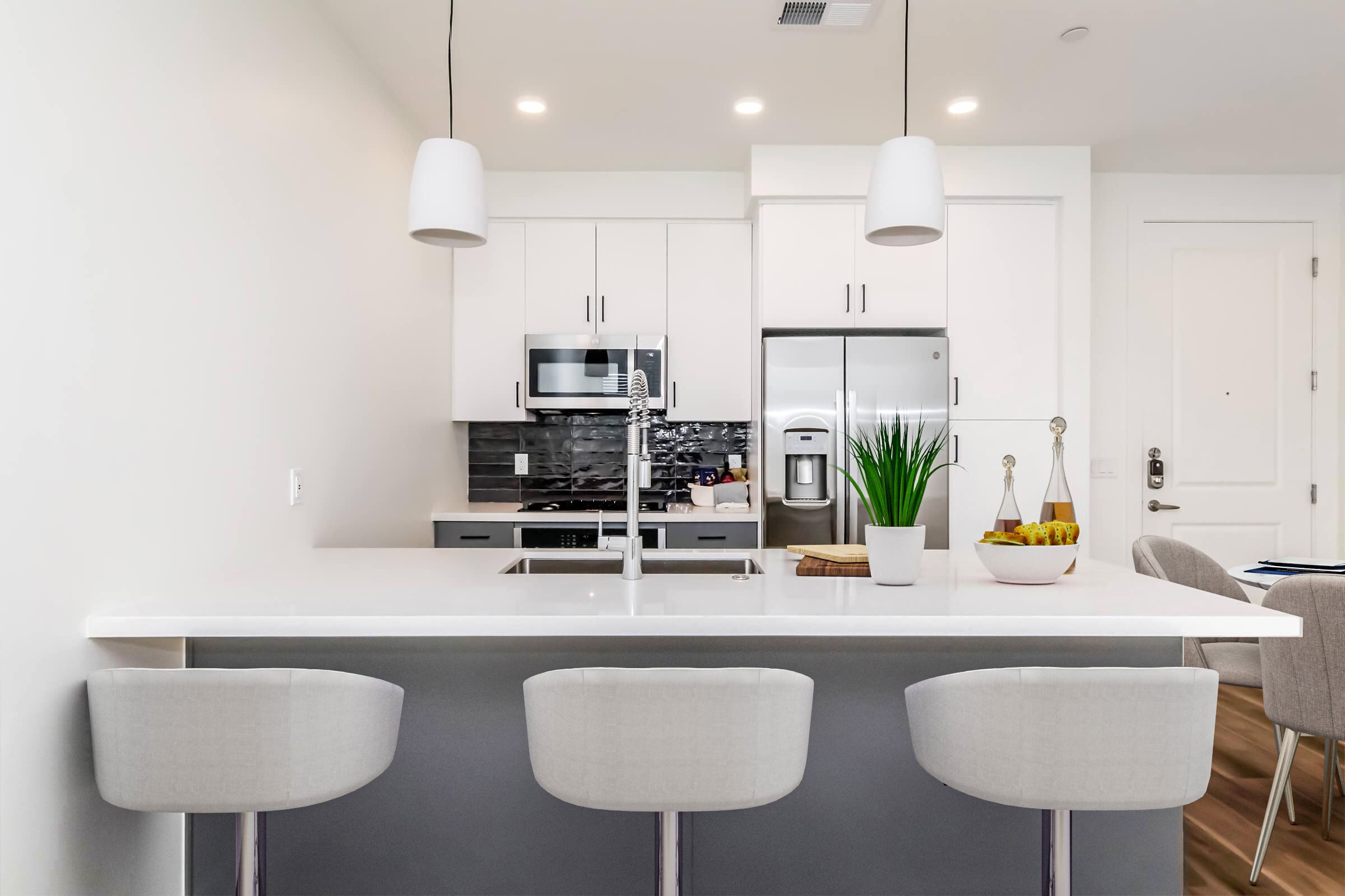 Modern kitchen with a quartz countertop and three light gray bar stools. Features include white cabinets, stainless steel appliances, a black and white tiled backsplash, and a potted plant on the counter. Bright lighting enhances the spacious and contemporary design. A dining area is visible in the background.